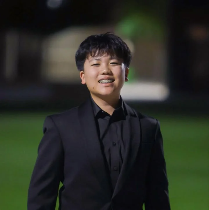 Young boy in a black suit and shirt standing outdoors at night, smiling.