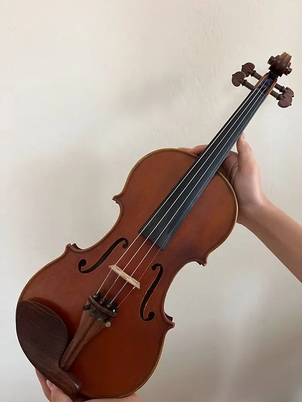 A person holding a wooden violin against a plain light-colored wall.