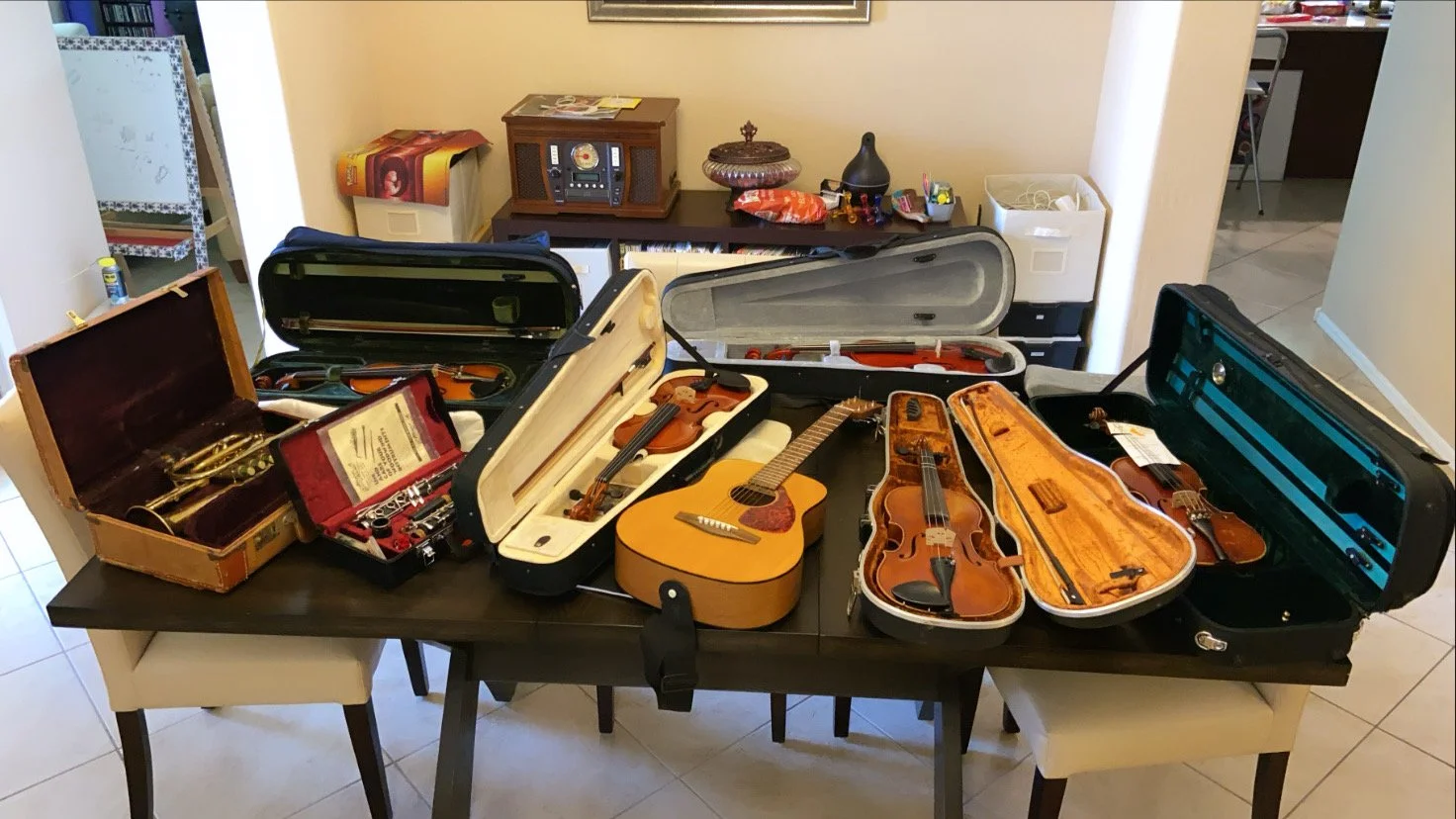 A dining table displaying several open and closed violins and guitars in their cases, with some instruments in hard cases and others in soft cases, in a room with a sideboard and various decorative items.