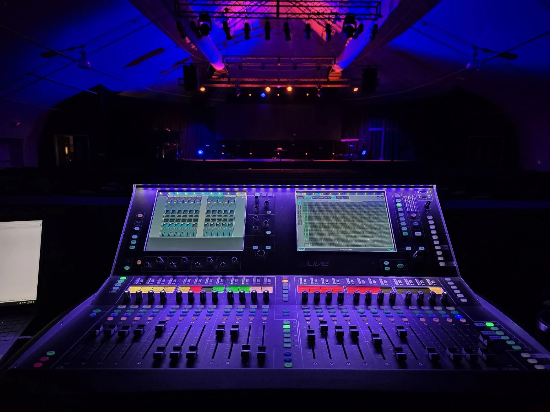 A digital audio mixing console with two screens, set up in a dimly lit stage or concert venue with colorful stage lighting.