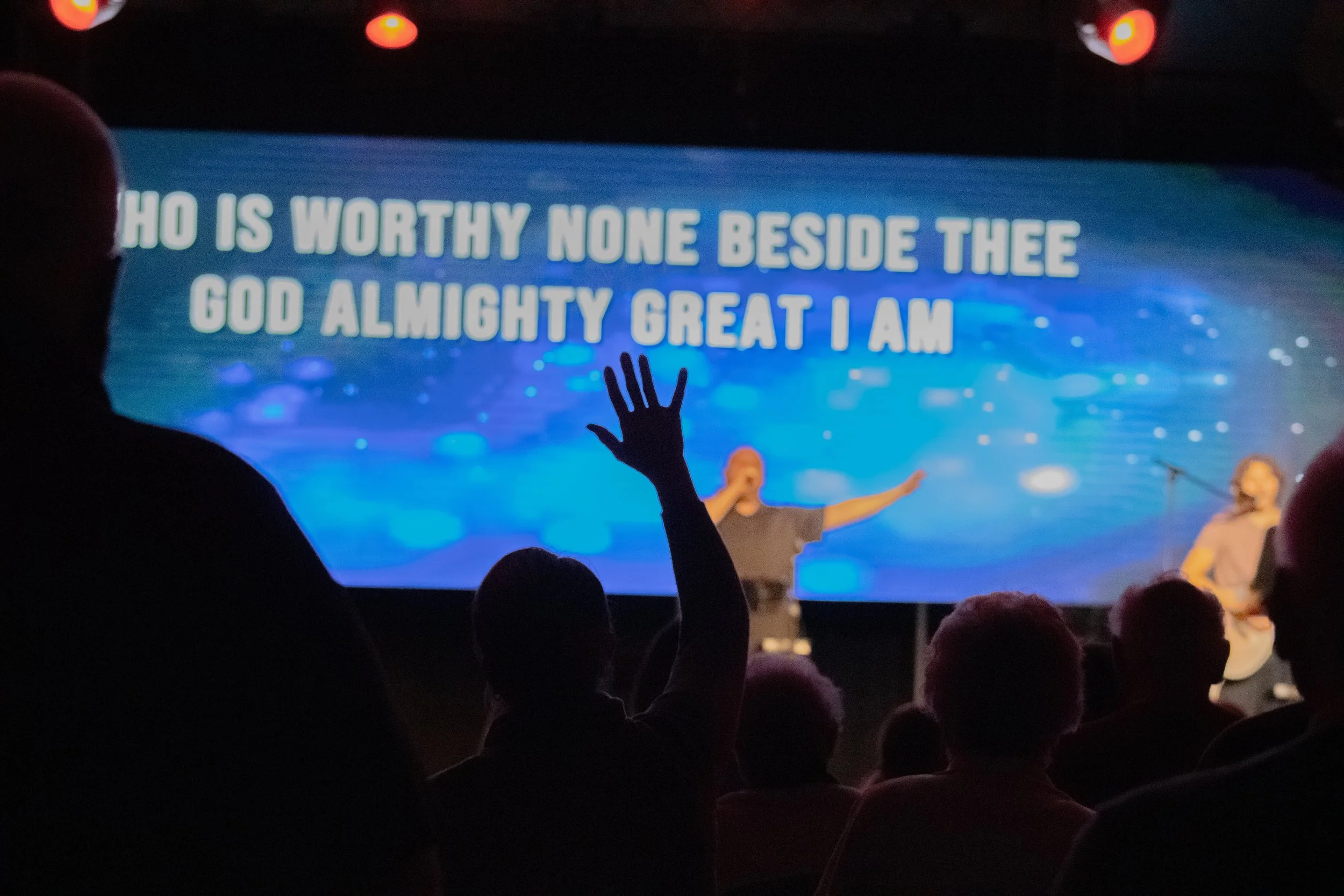 A group of people attending a worship service or concert. One person in the audience has their hand raised. On stage, there is a man with outstretched arms and a woman with a guitar. A large screen behind displays the lyrics: "WHO IS WORTHY NONE BESIDE THEE GOD ALMIGHTY GREAT I AM." The scene is dimly lit with some red stage lights.