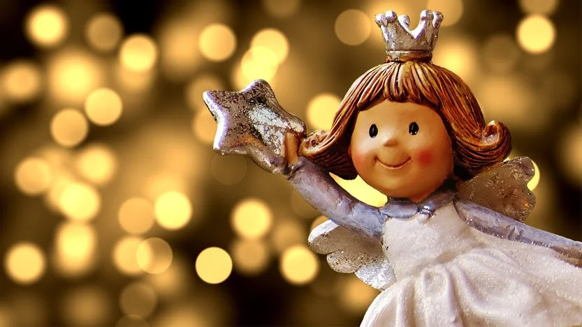 Decorative angel figurine wearing a crown, holding a star-shaped wand, with a background of blurred warm lights.