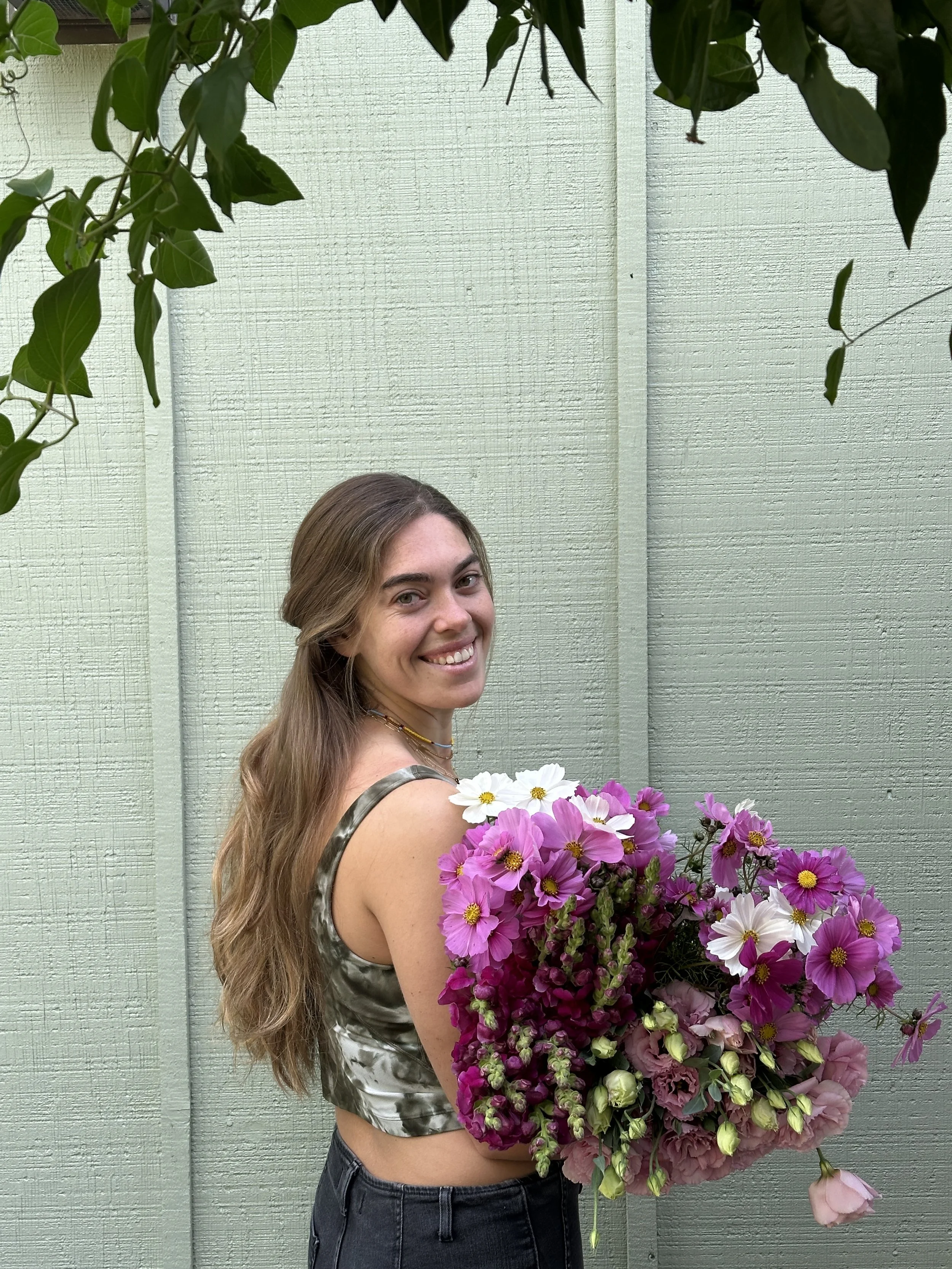 A young woman with long, wavy brown hair is smiling and holding a large bouquet of pink and white flowers. She is wearing a sleeveless, patterned crop top and dark jeans, standing against a light green wooden wall with greenery overhead.
