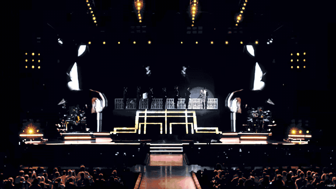 Stage with illuminated stairs and performers, large screens displaying black and white images, audience in foreground, concert or performance setting.