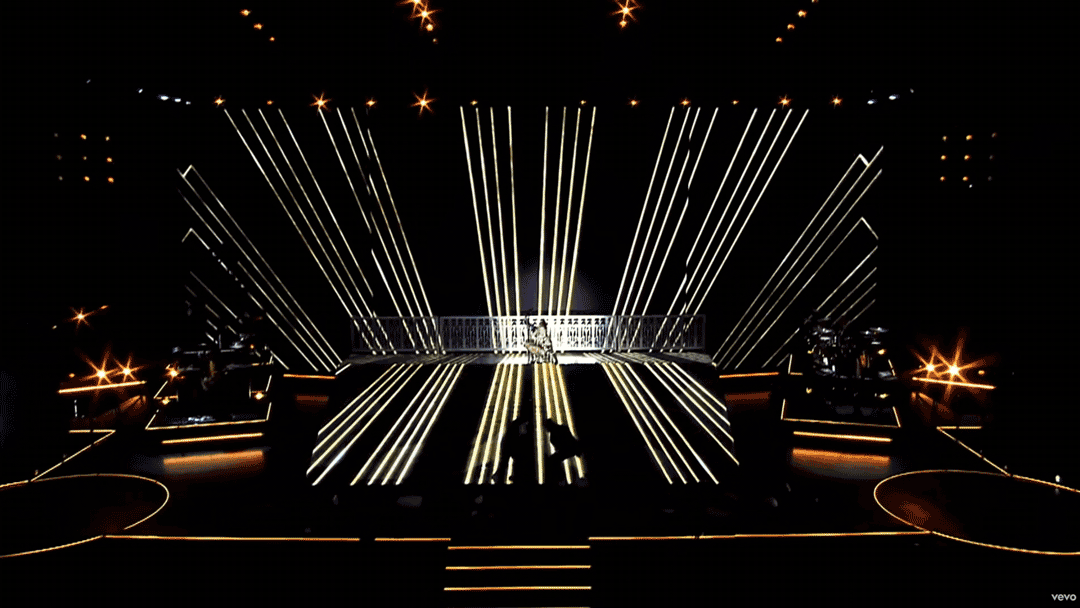 Stage with dynamic vertical and horizontal light streaks, spotlight effects, and silhouettes of performers or dancers, creating a futuristic visual display.