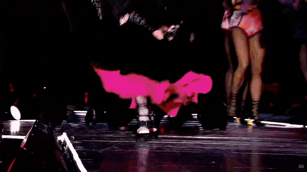 A performer on stage wearing a black outfit with a pink sash bending forward, with other performers in the background.