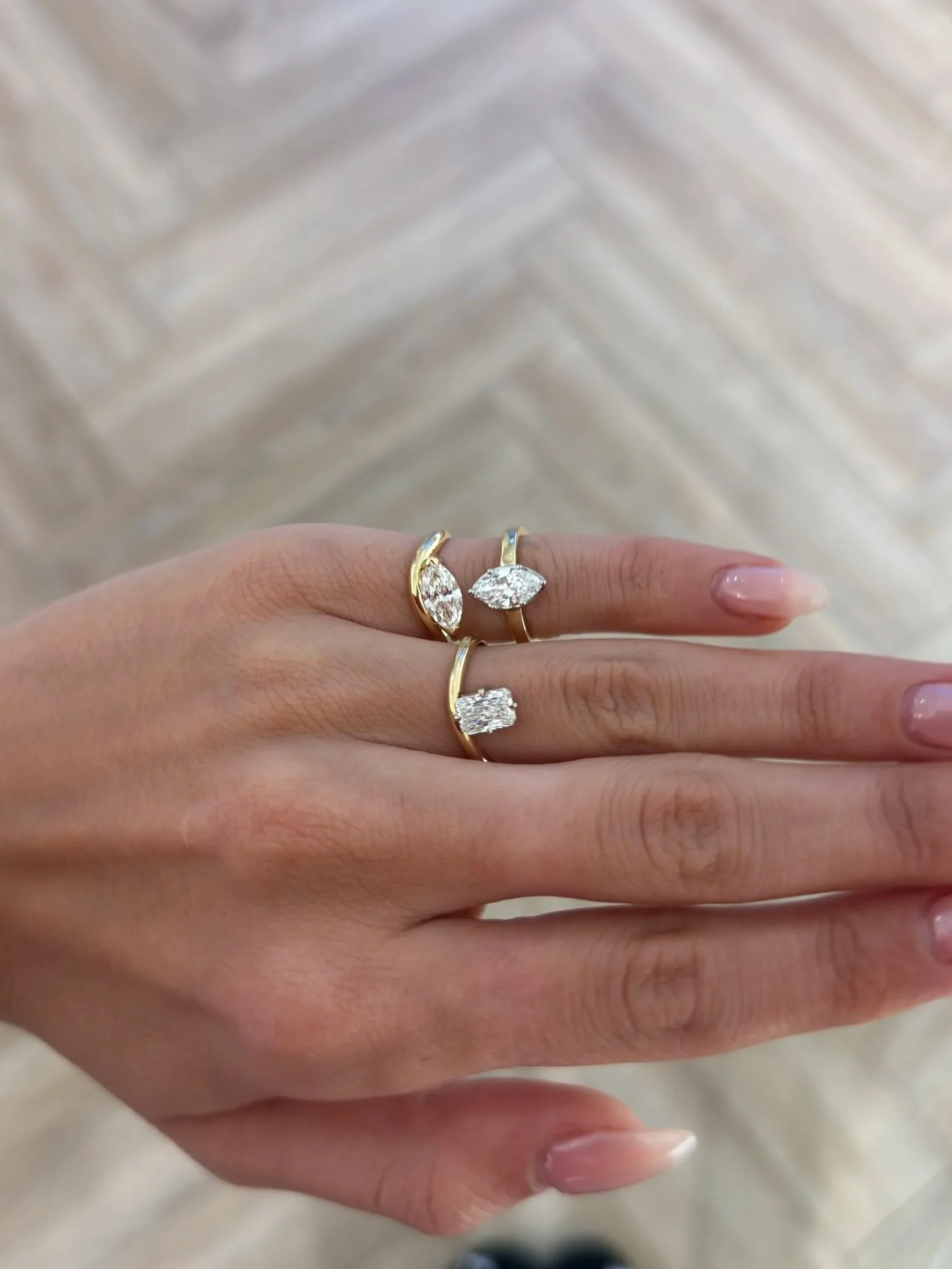 A close-up of a hand wearing multiple gold rings with large diamond stones, set against a light, textured wooden background.