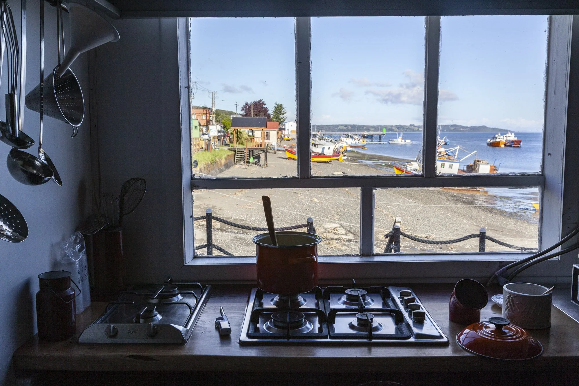 Vista de la cocina al interior, con estufa de dos quemadores con un cazo rojo y una cuchara, y otros utensilios de cocina en la encimera, mirando hacia un puerto con barcos en el agua y edificios frente a la playa.