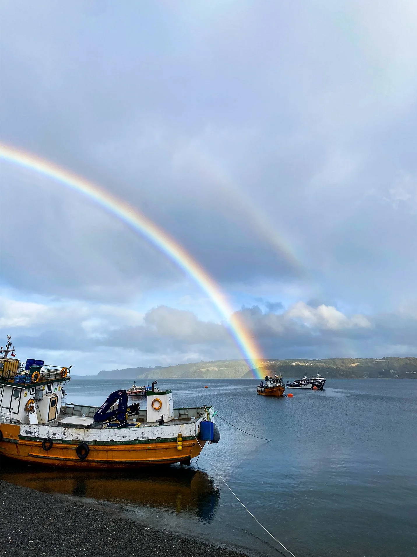 Dos arcos de arcoíris sobre barcos en un puerto con cielo nublado y horizonte con colinas.