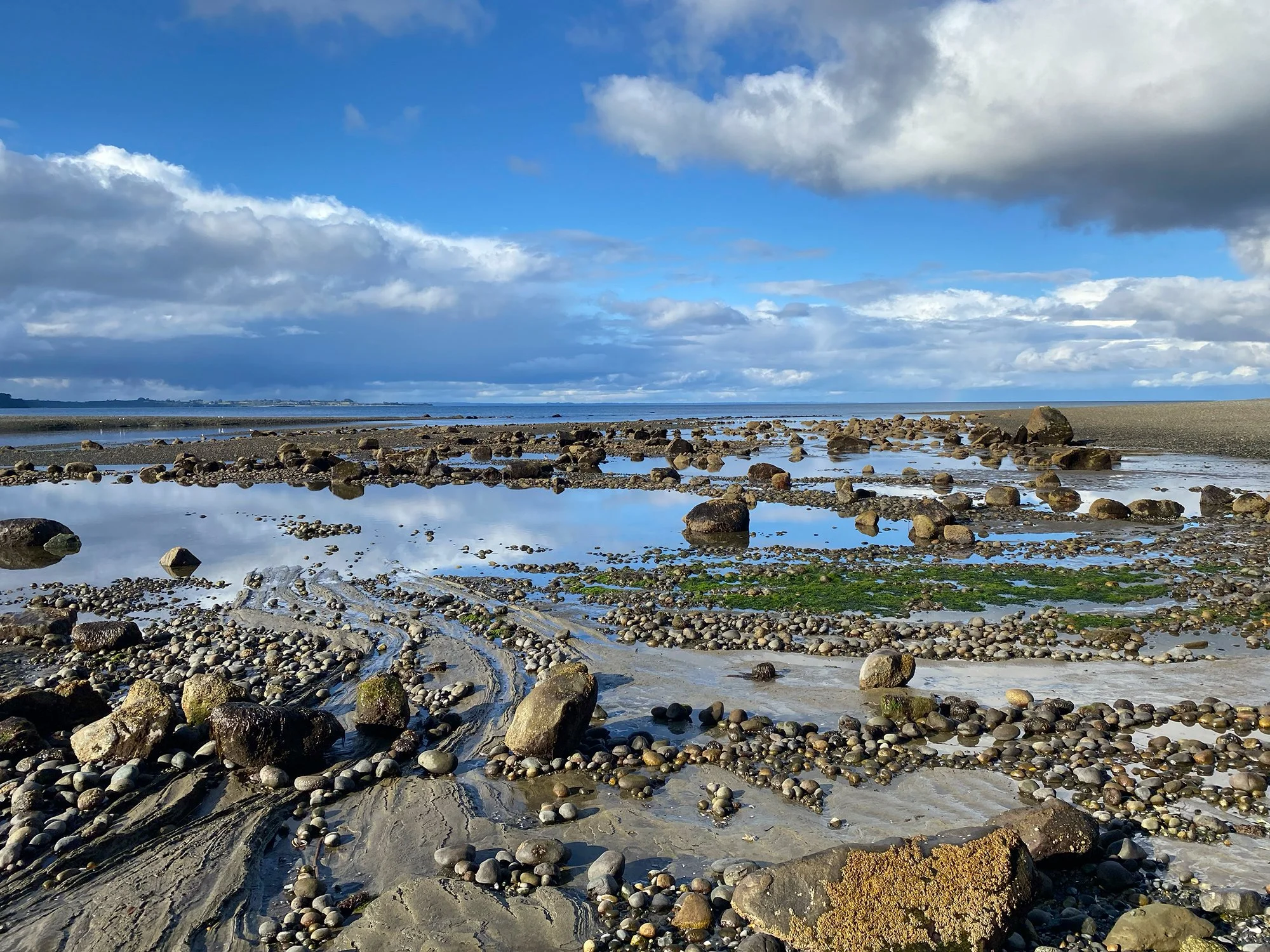 Costa rocosa con piedras y charcos de agua, cielo nuboso y algunos rayos de sol, mar en la distancia.