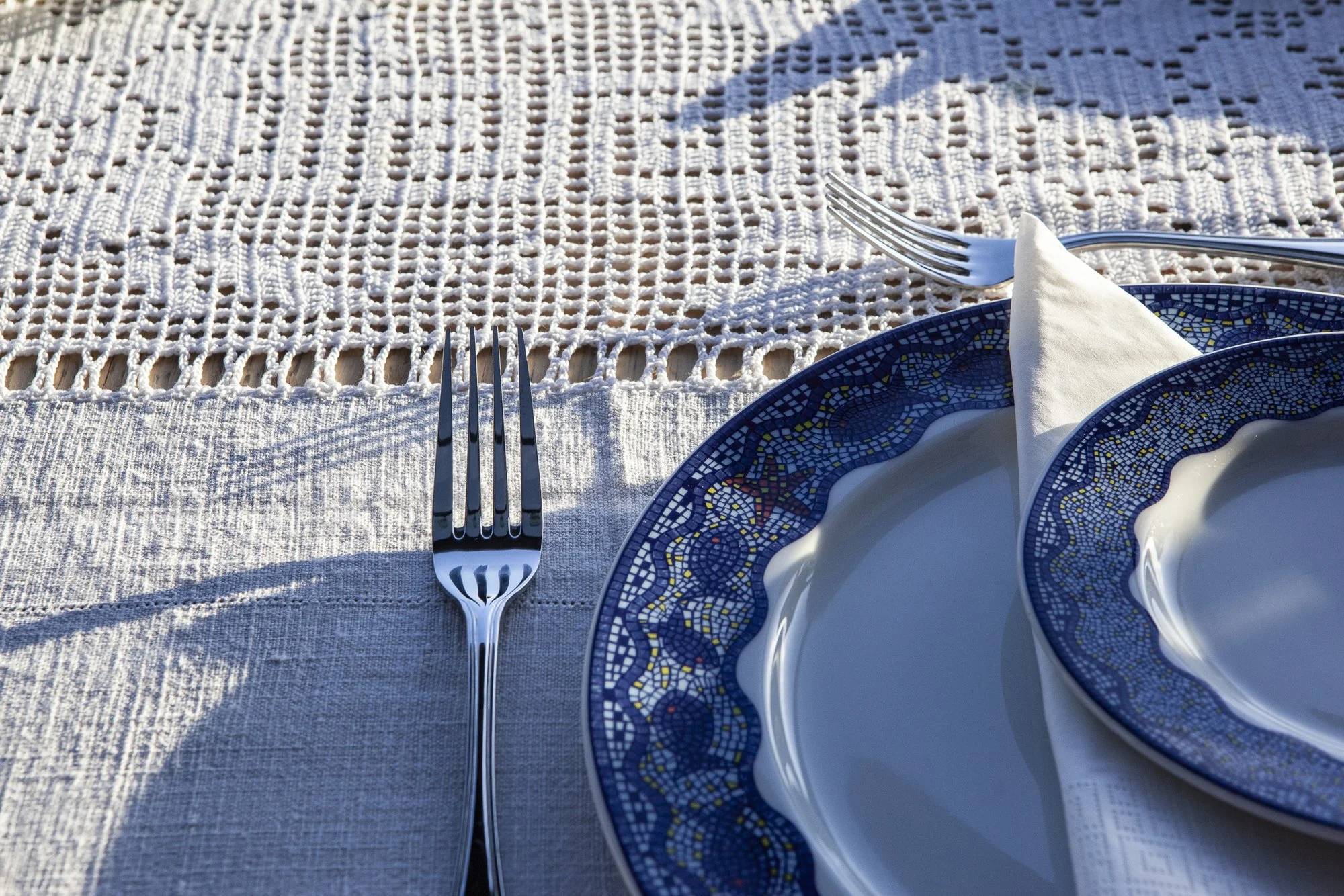 Configuración de mesa con platos y cubiertos en una mesa al aire libre, con mantel y caminos de mesa de tela, en un entorno soleado.