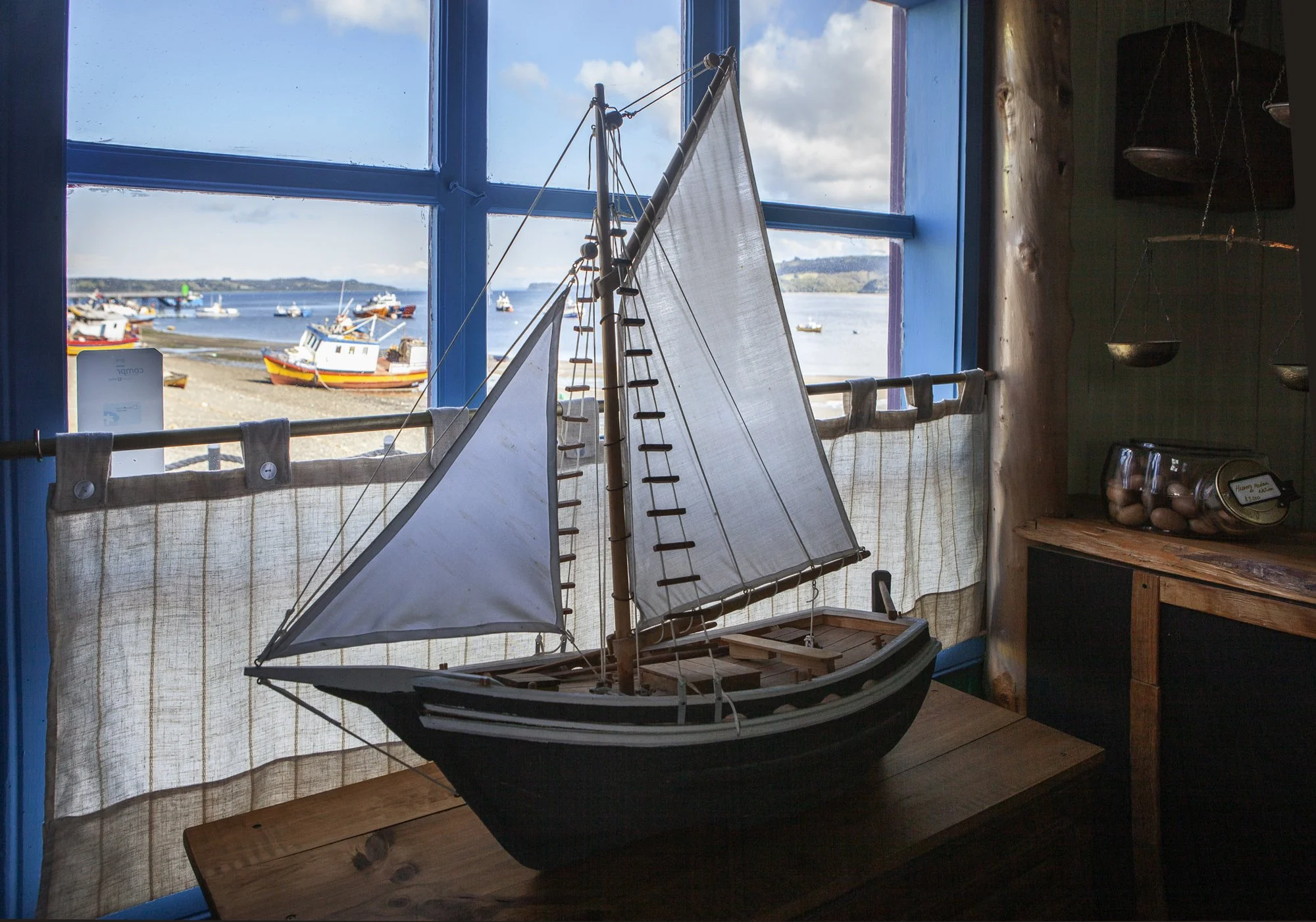 Modelado de barco de vela en una habitación, con vista a un puerto por la ventana.