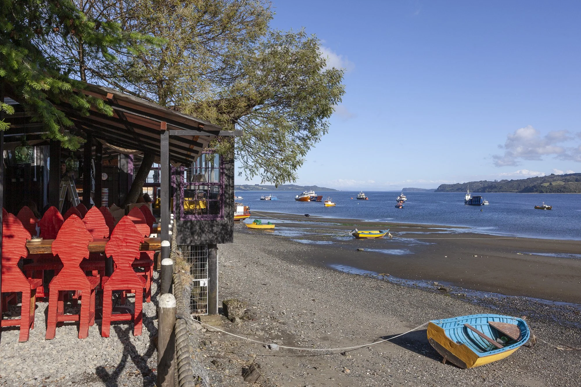 Visto del puerto con barcos en el agua, algunos en la orilla, y un restaurante con mesas y sillas rojas a la izquierda, junto a árboles, en un día soleado con cielo despejado.