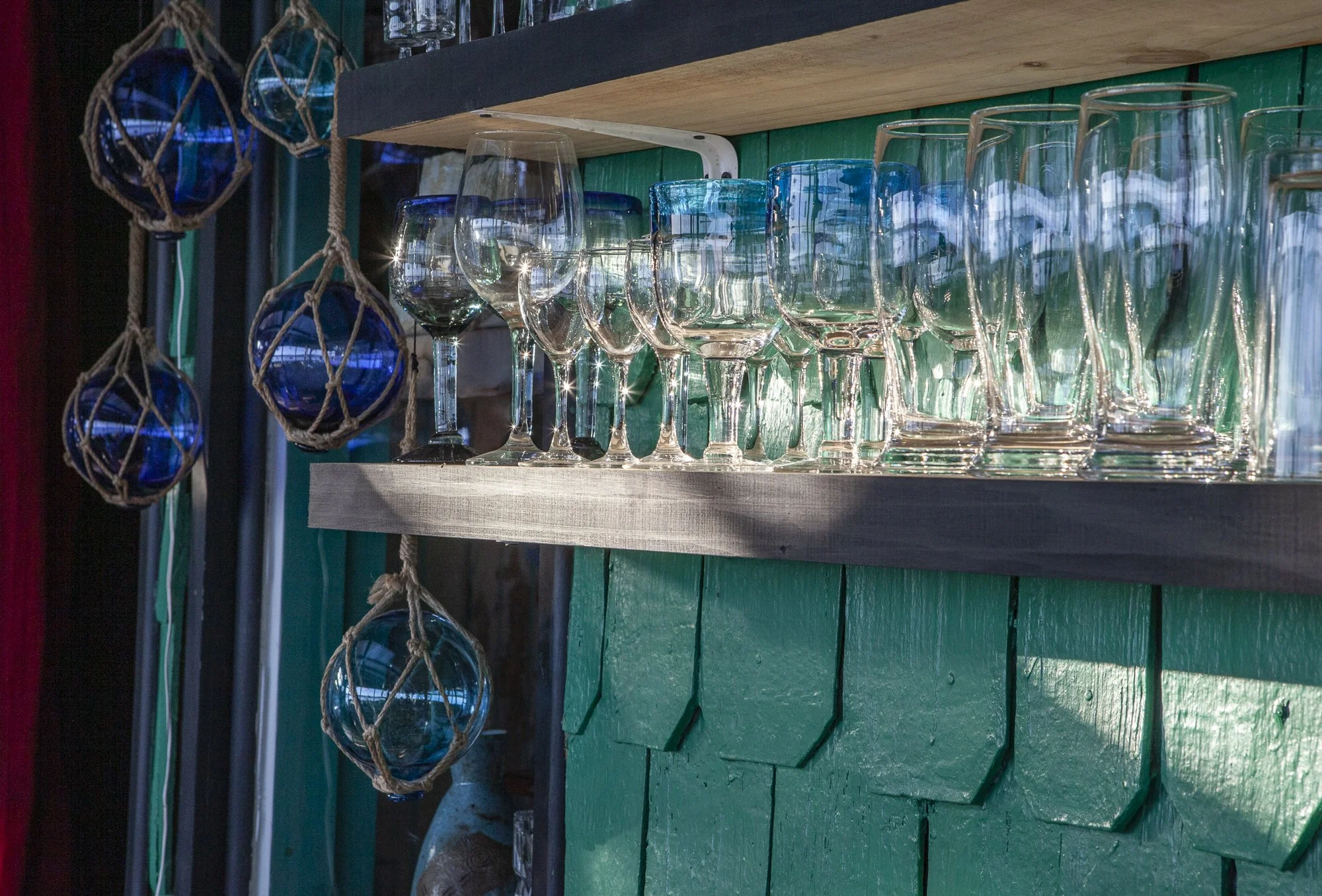 Vasos y copas de cristal en una estantería de madera, con decoraciones de bolas de vidrio enredadas con cuerda colgadas a la izquierda.