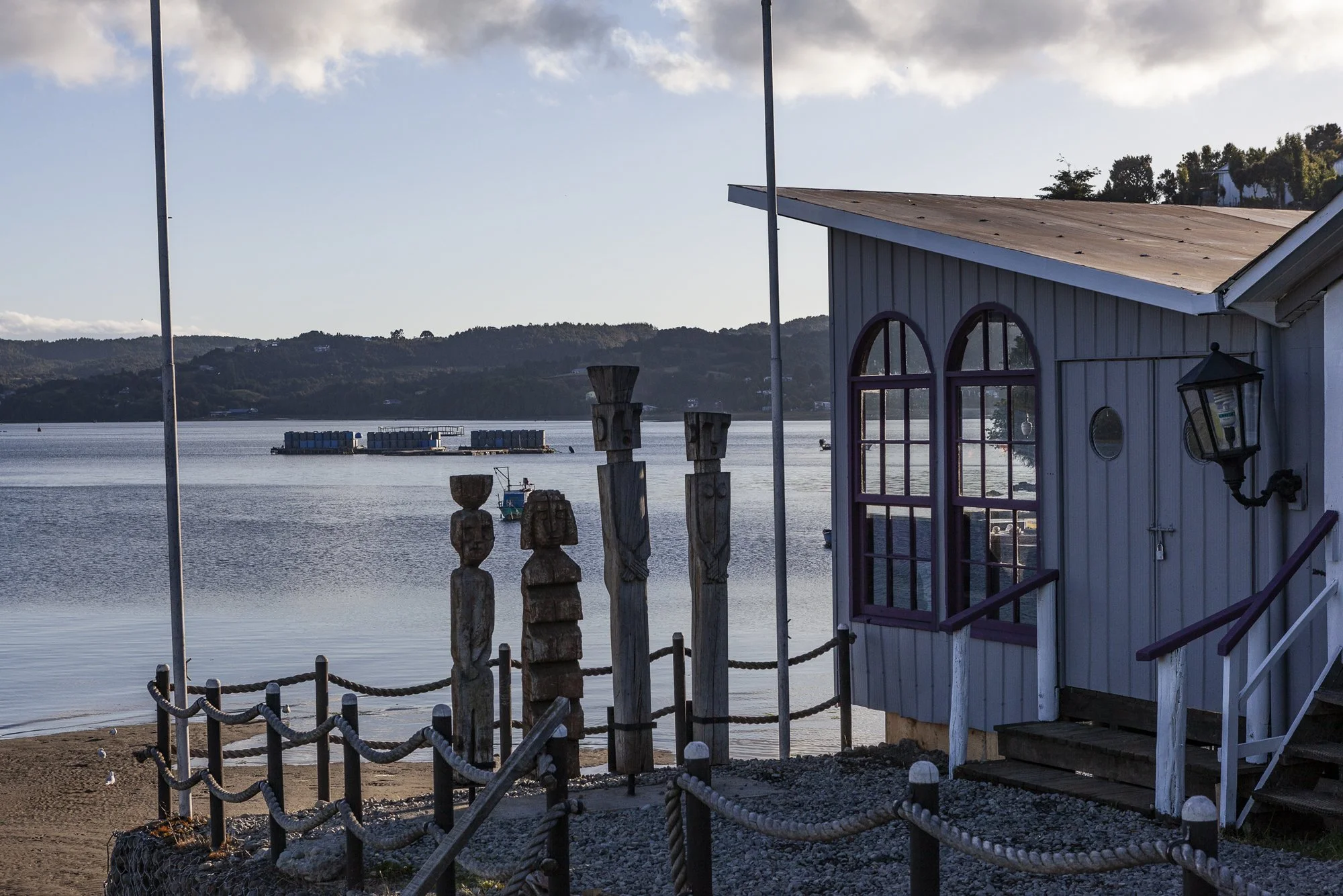 Casita junto al agua con lámpara de pared, barandilla de cuerdas, postres tallados en madera, y vista del río con barcos y montañas al fondo.