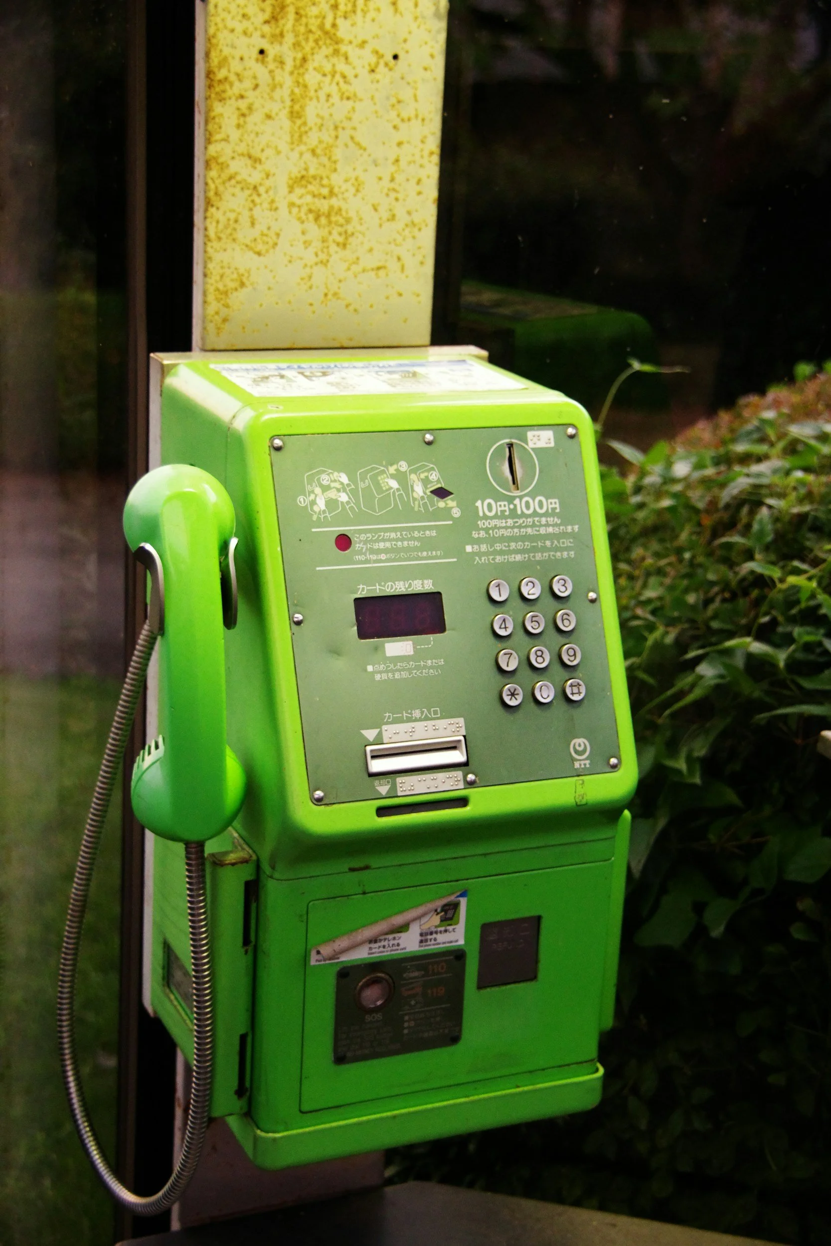 Green public telephone kiosk in Japan with a handset attached to the side and a keypad, placed outside near bushes.