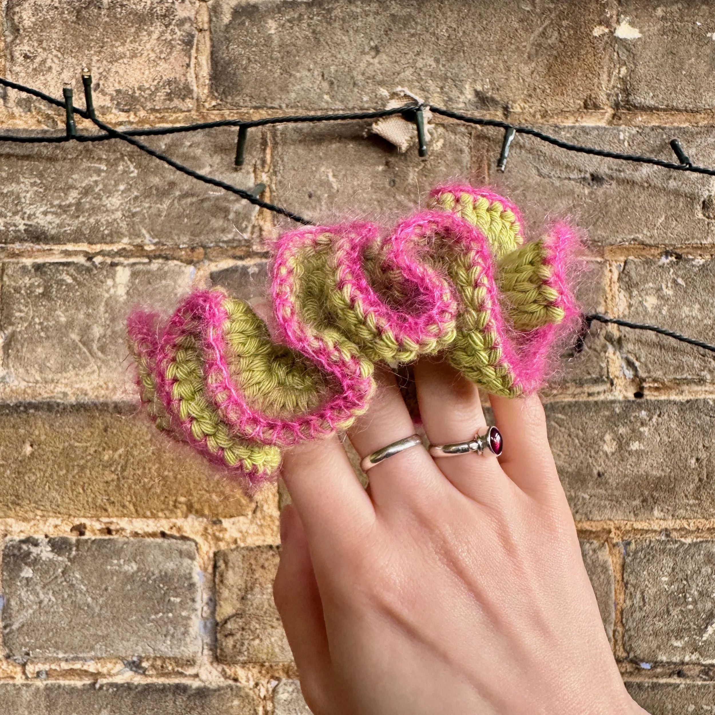 A bright green cotton crochet ruffle scrunchie with a pink mohair trim being held to show the shape