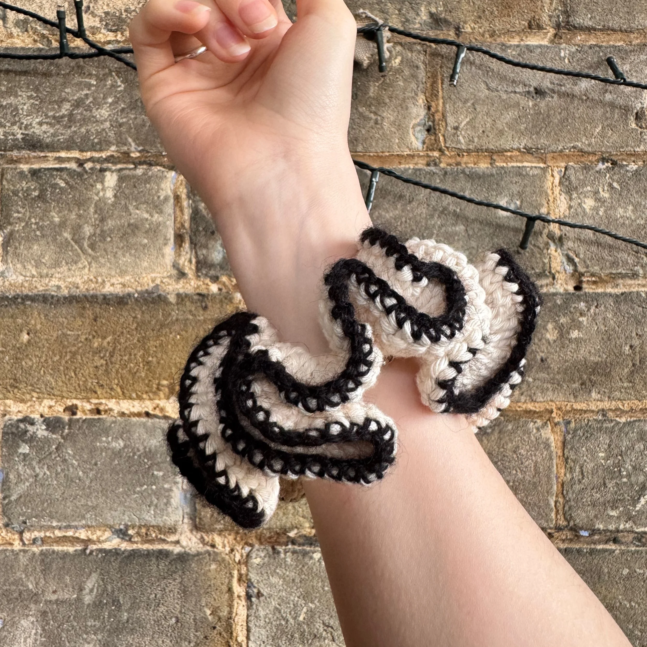 A cream cotton crochet ruffle scrunchie with a black alpaca trim being worn on a wrist