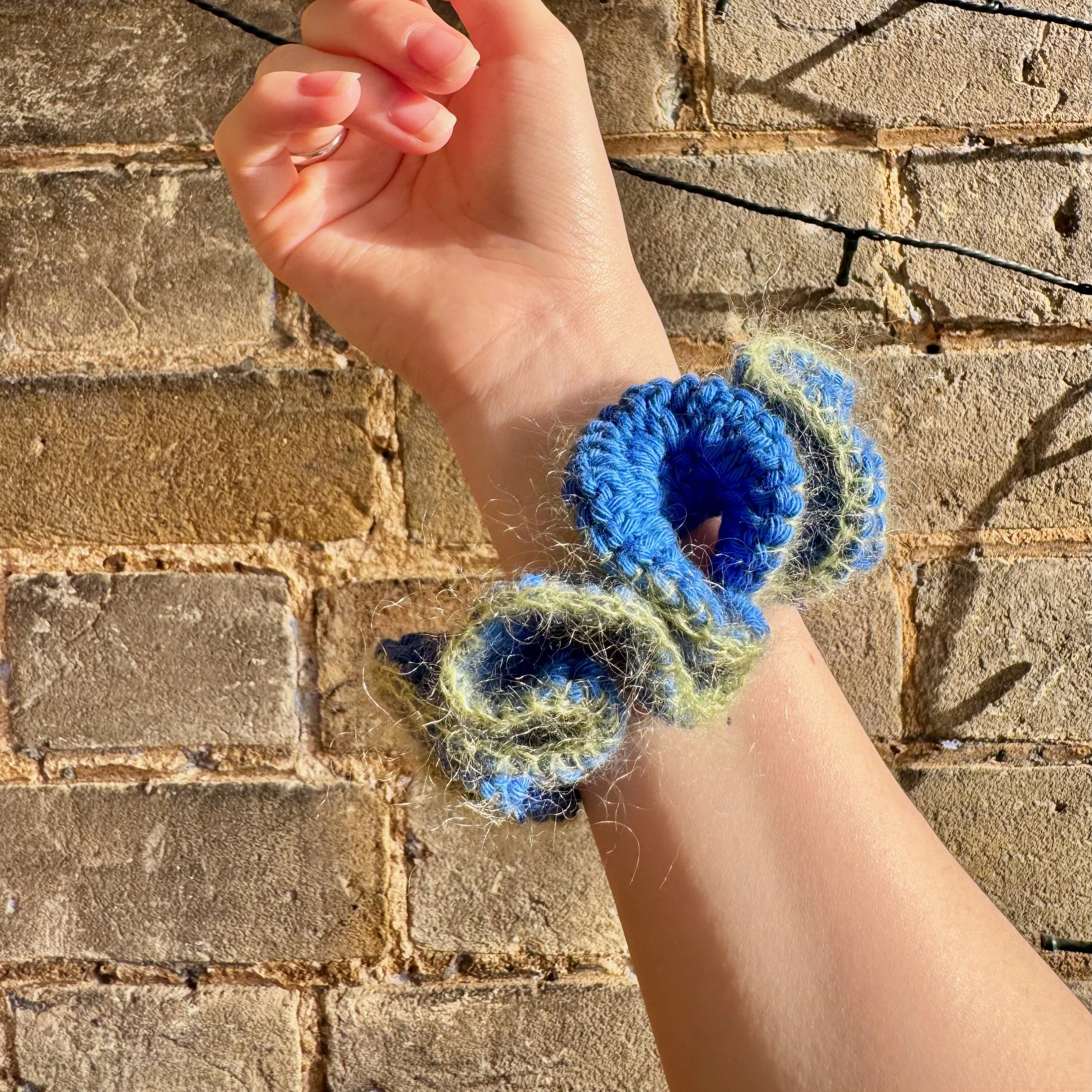 A bright blue cotton crochet ruffle scrunchie with a green mohair trim being worn on a wrist