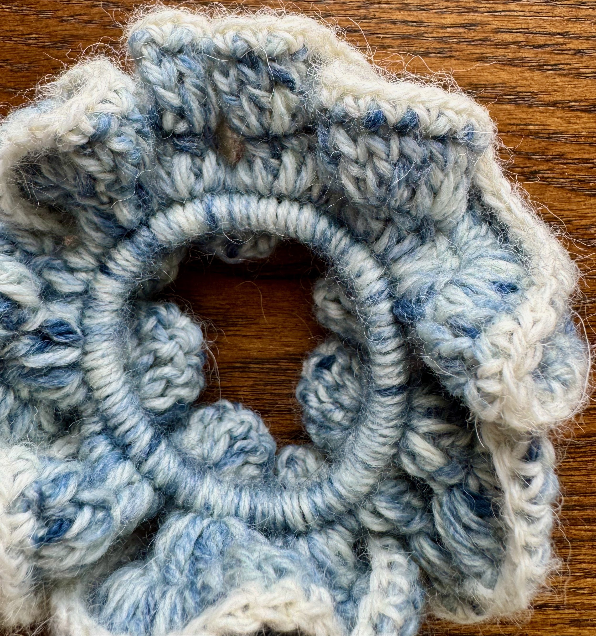A multi blue woollen crochet ruffle scrunchie that looks like denim, with a white alpaca trim laying on a flat surface zoomed in to show details