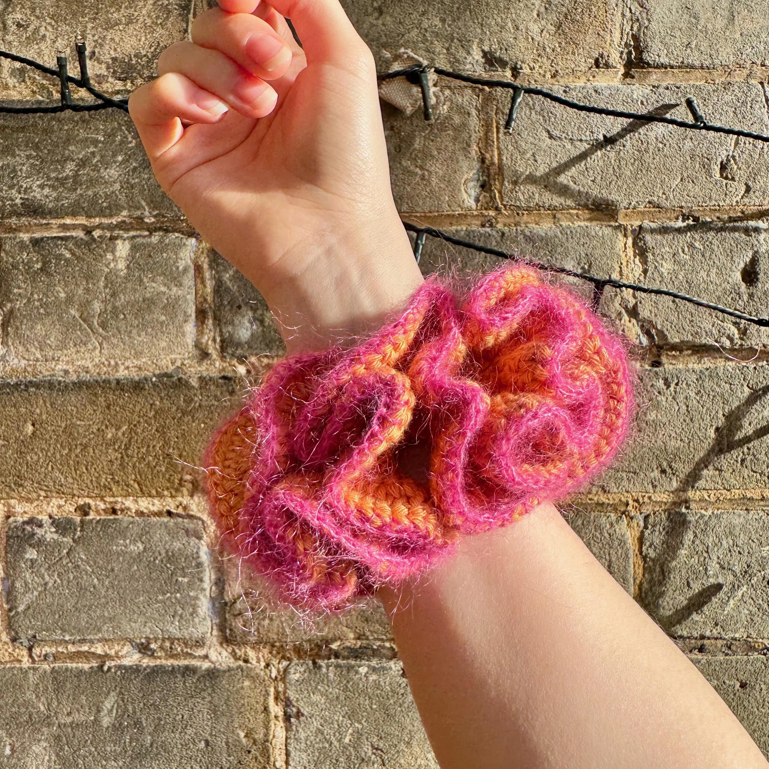An orange crochet ruffle scrunchie with a pink mohair trim being worn on a wrist