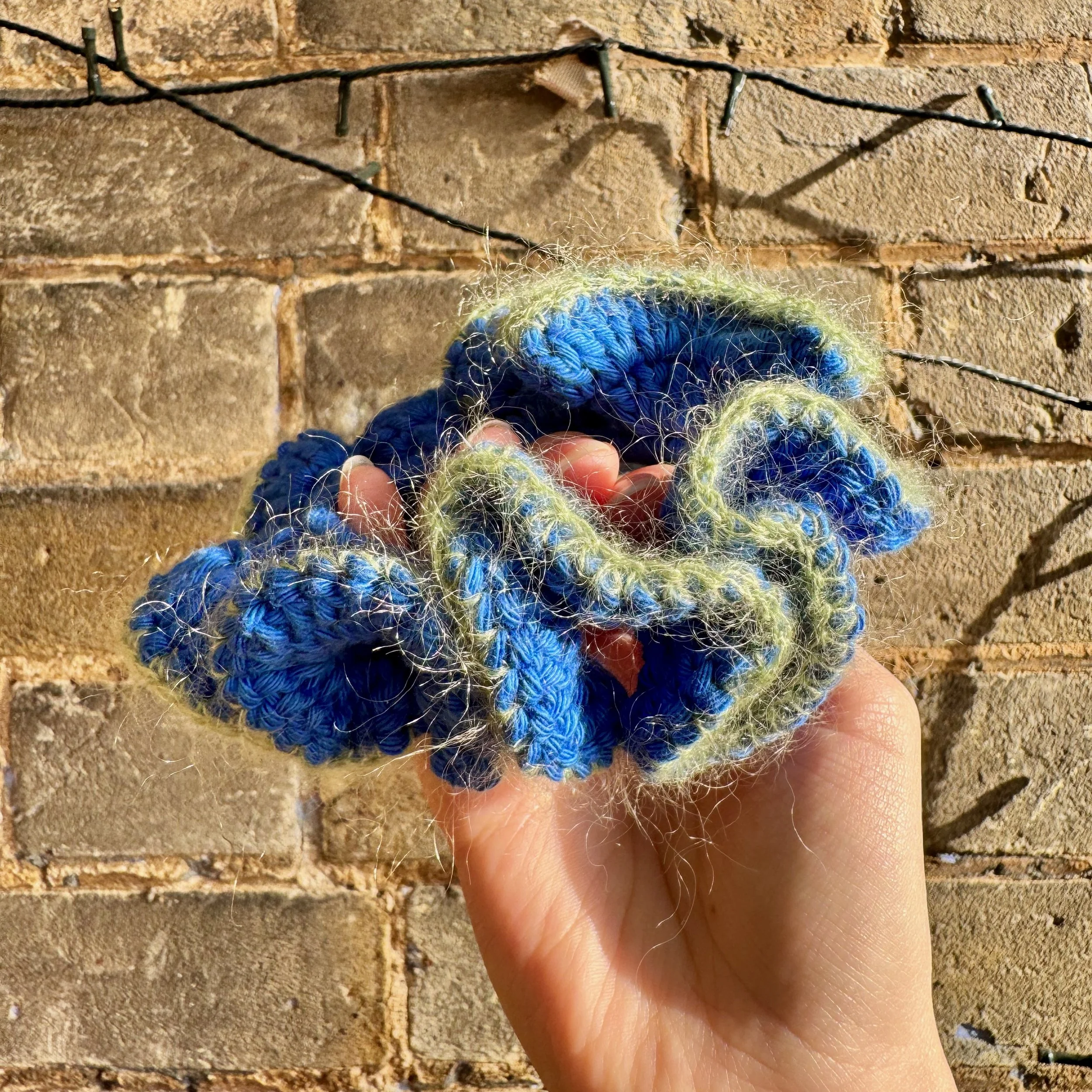 A bright blue cotton crochet ruffle scrunchie with a green mohair trim being held to show the ruffles