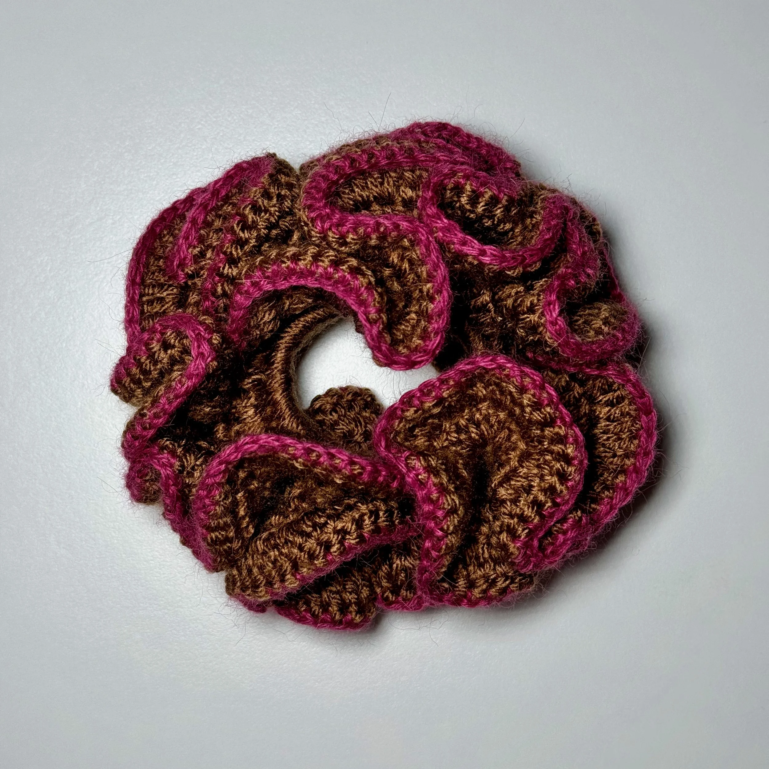 A brown crochet ruffle scrunchie with a pink alpaca trim laying flat on a surface