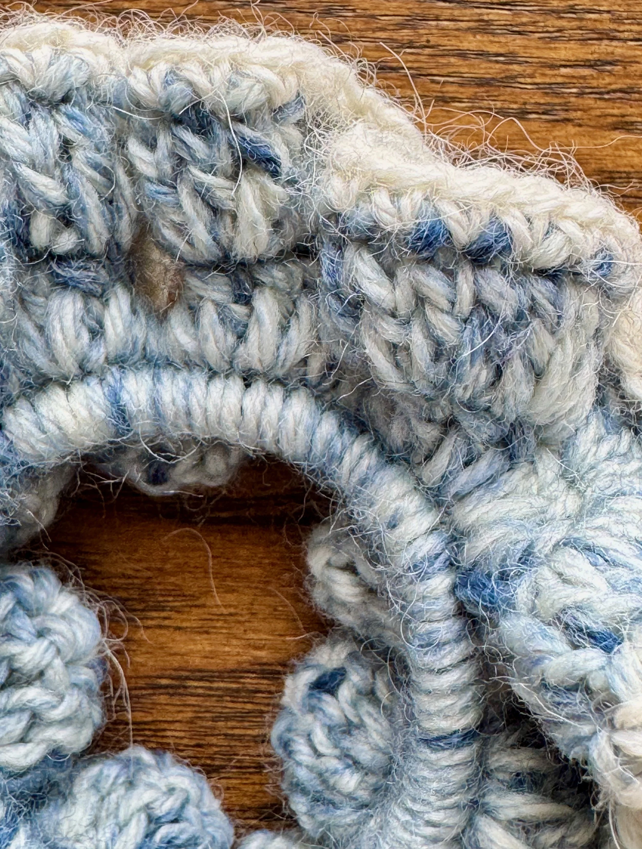 A multi blue woollen crochet ruffle scrunchie that looks like denim, with a white alpaca trim laying on a flat surface zoomed in to show details