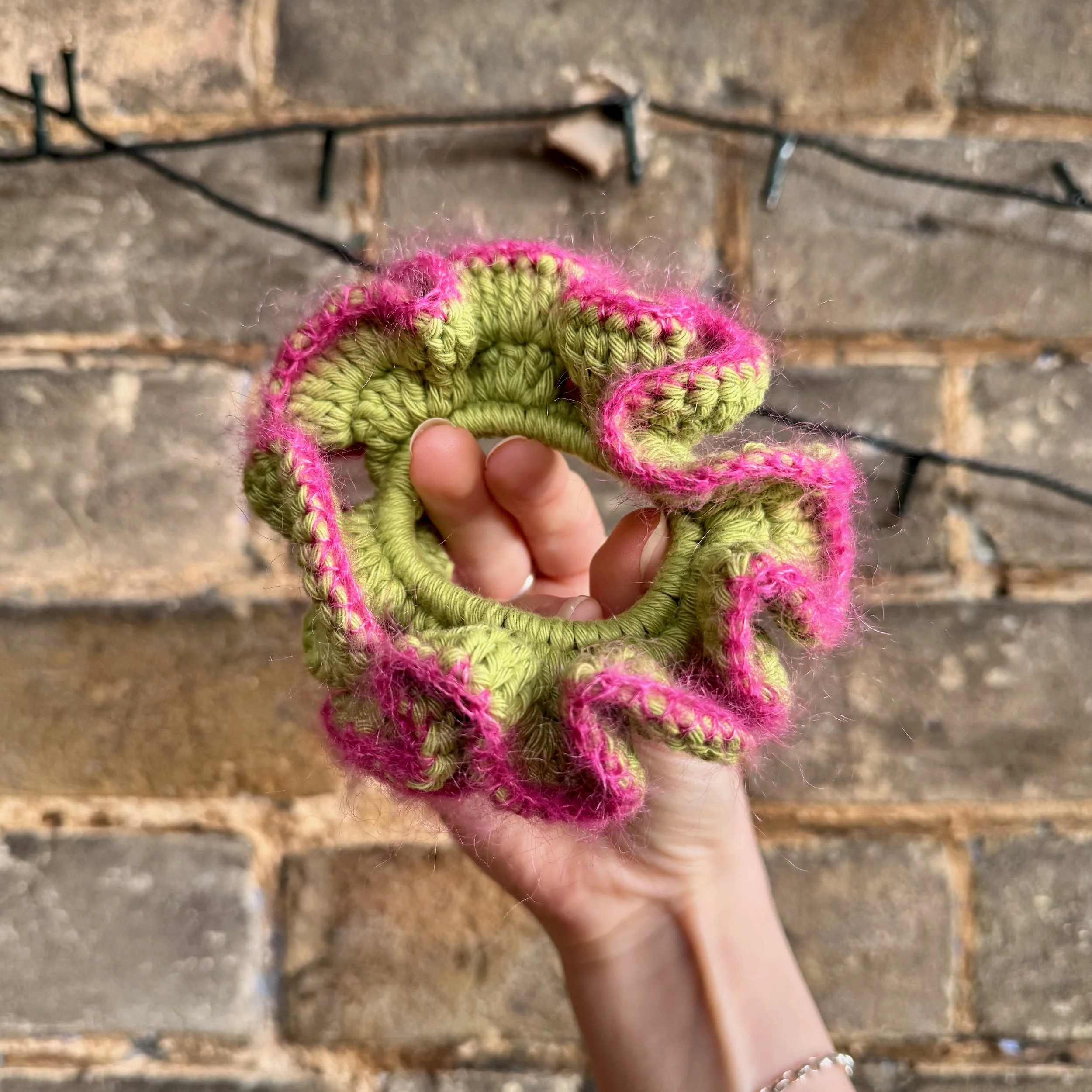 A bright green cotton crochet ruffle scrunchie with a pink mohair trim being held to show the shape