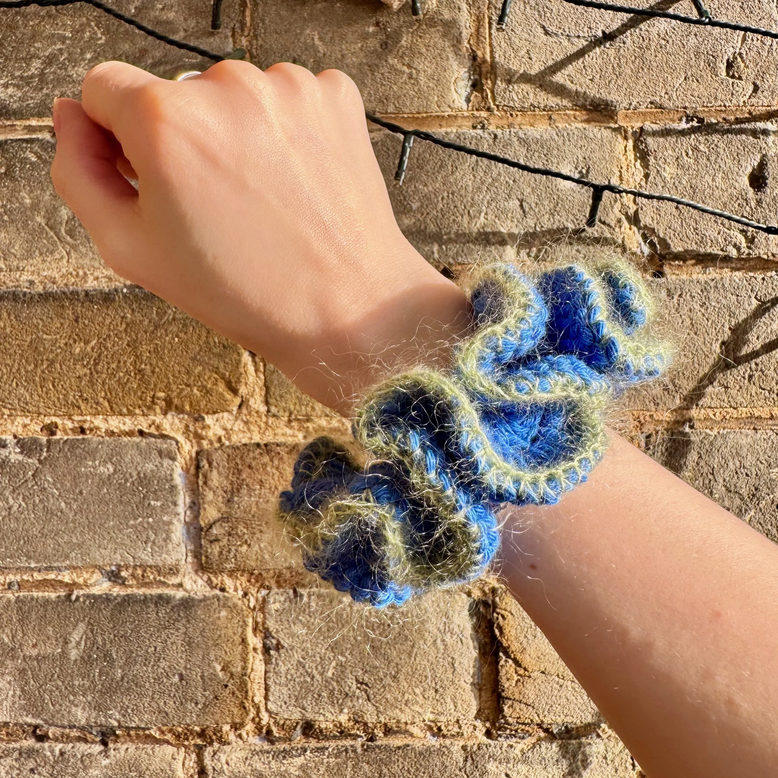 A bright blue cotton crochet ruffle scrunchie with a green mohair trim being worn on a wrist