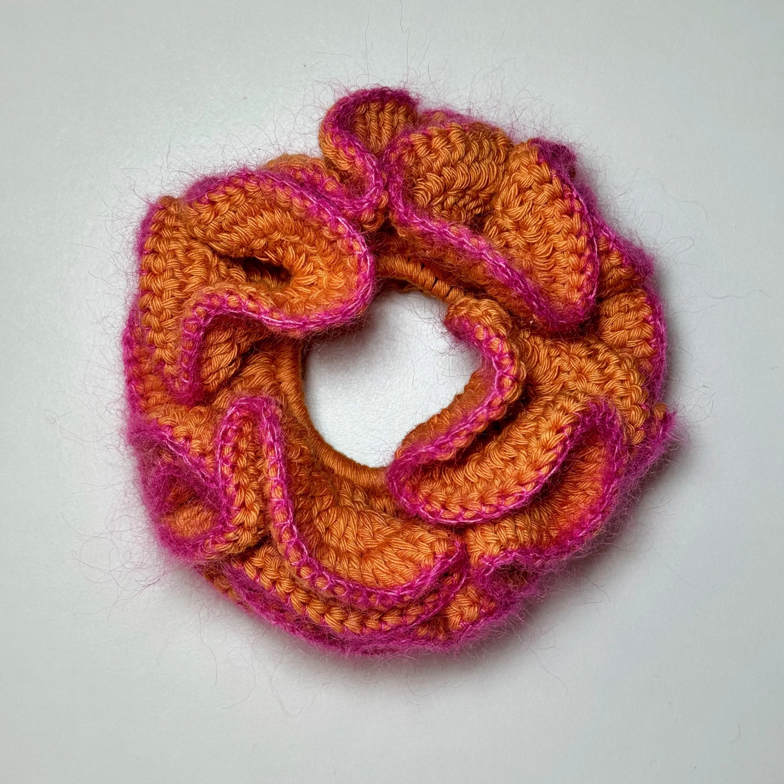 An orange crochet ruffle scrunchie with a pink mohair trim laying flat on a surface