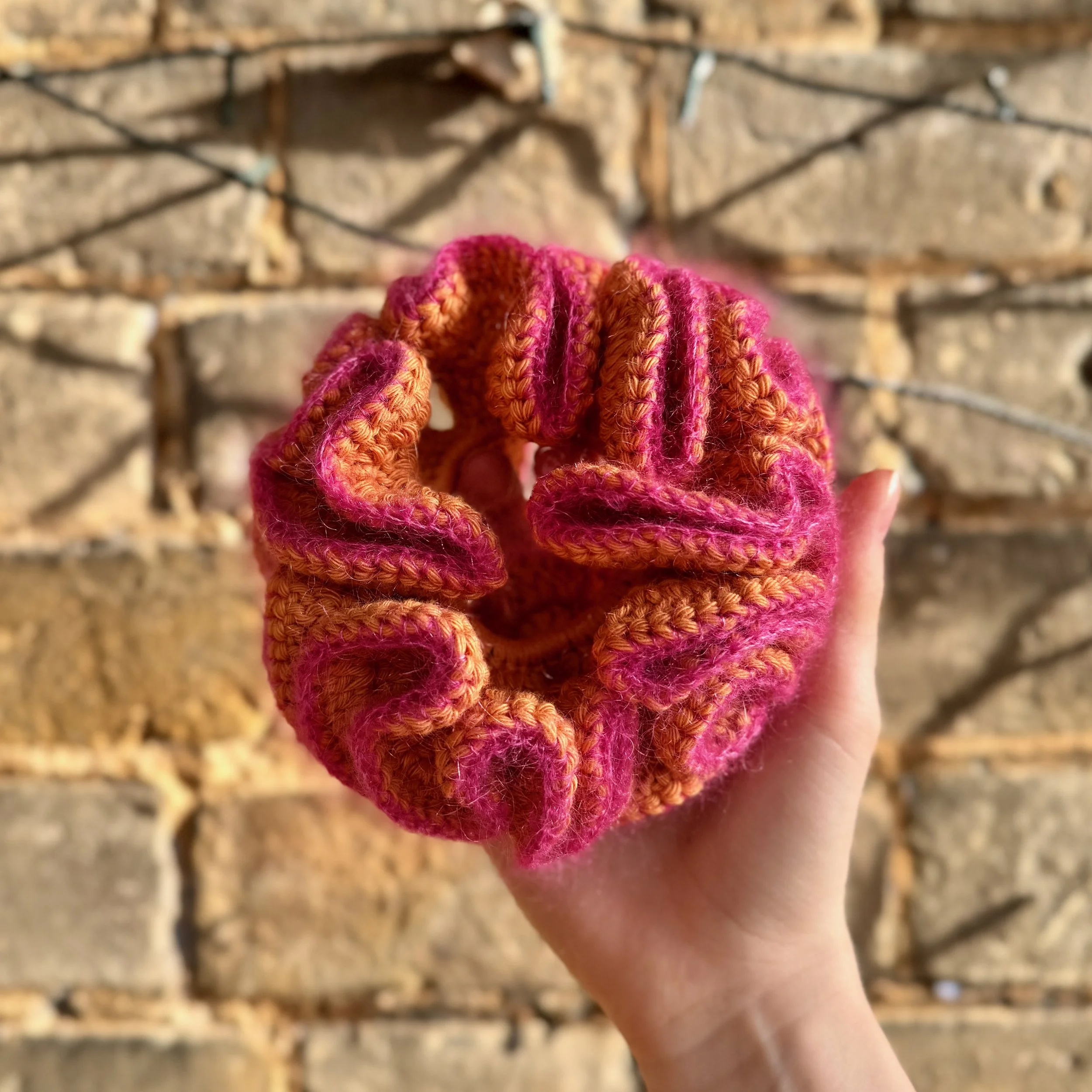 An orange crochet ruffle scrunchie made from cotton with a pink mohair trim being held to show the ruffles.