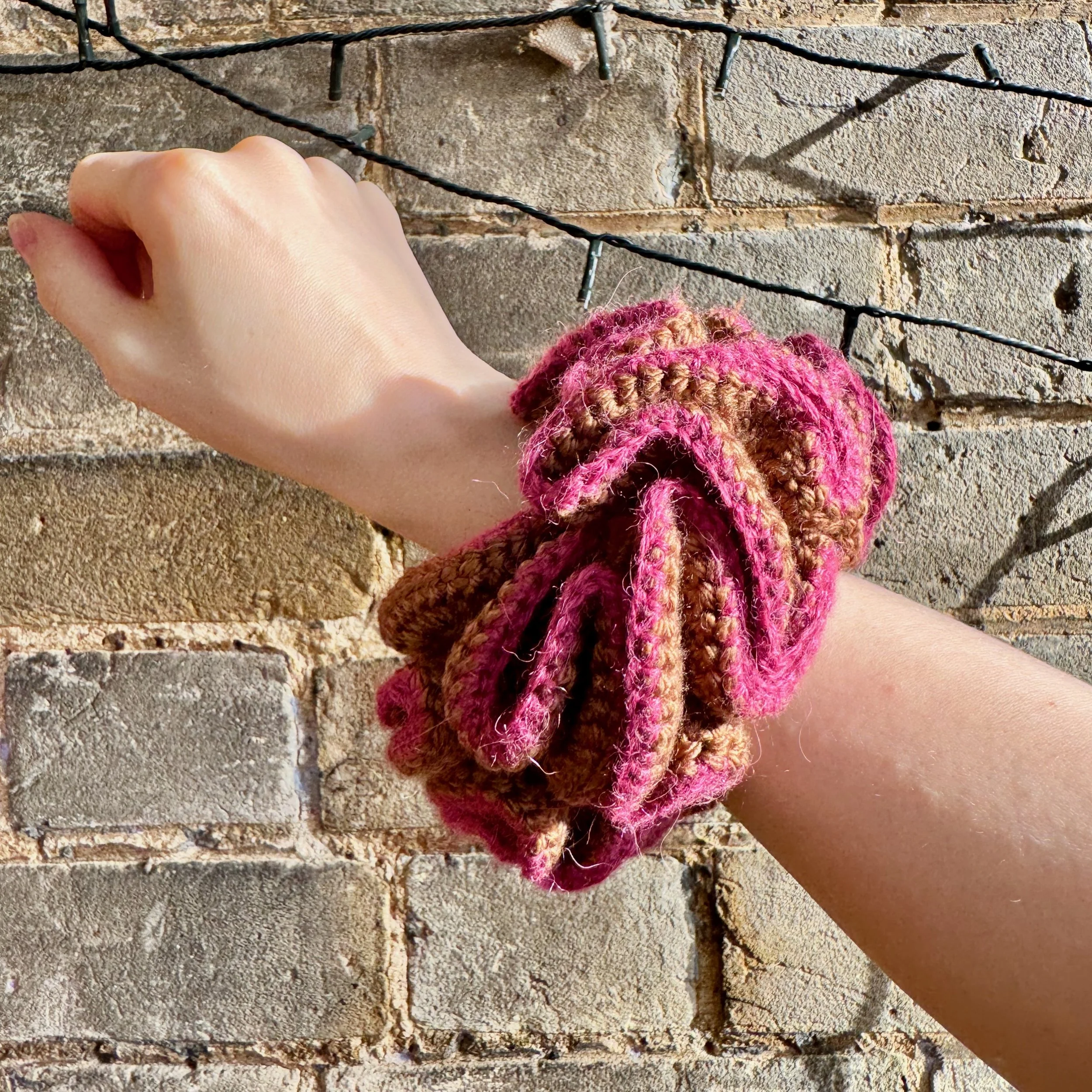 A brown crochet ruffle scrunchie with a pink alpaca trim being worn on a wrist