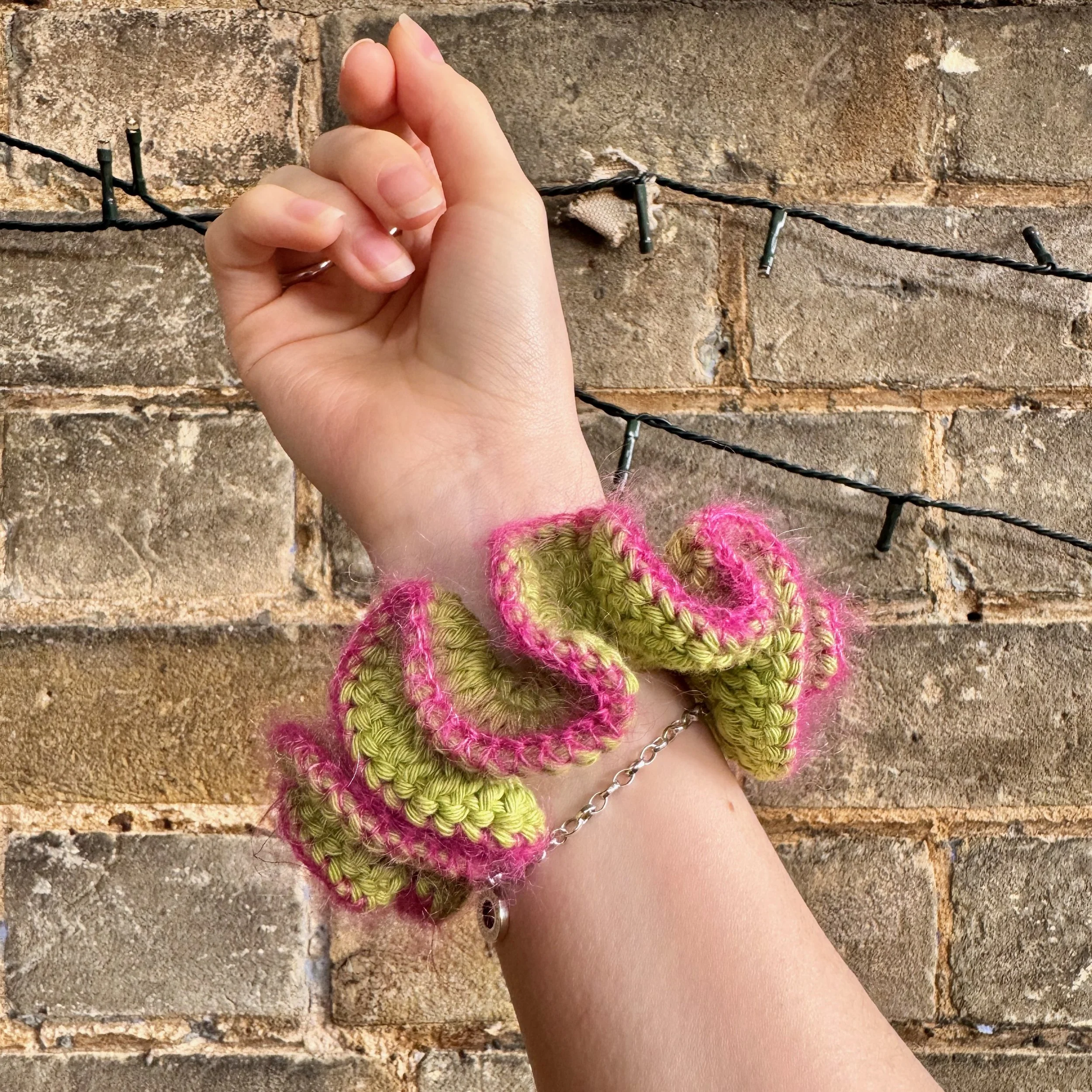 A bright green cotton crochet ruffle scrunchie with a pink mohair trim being worn on a wrist