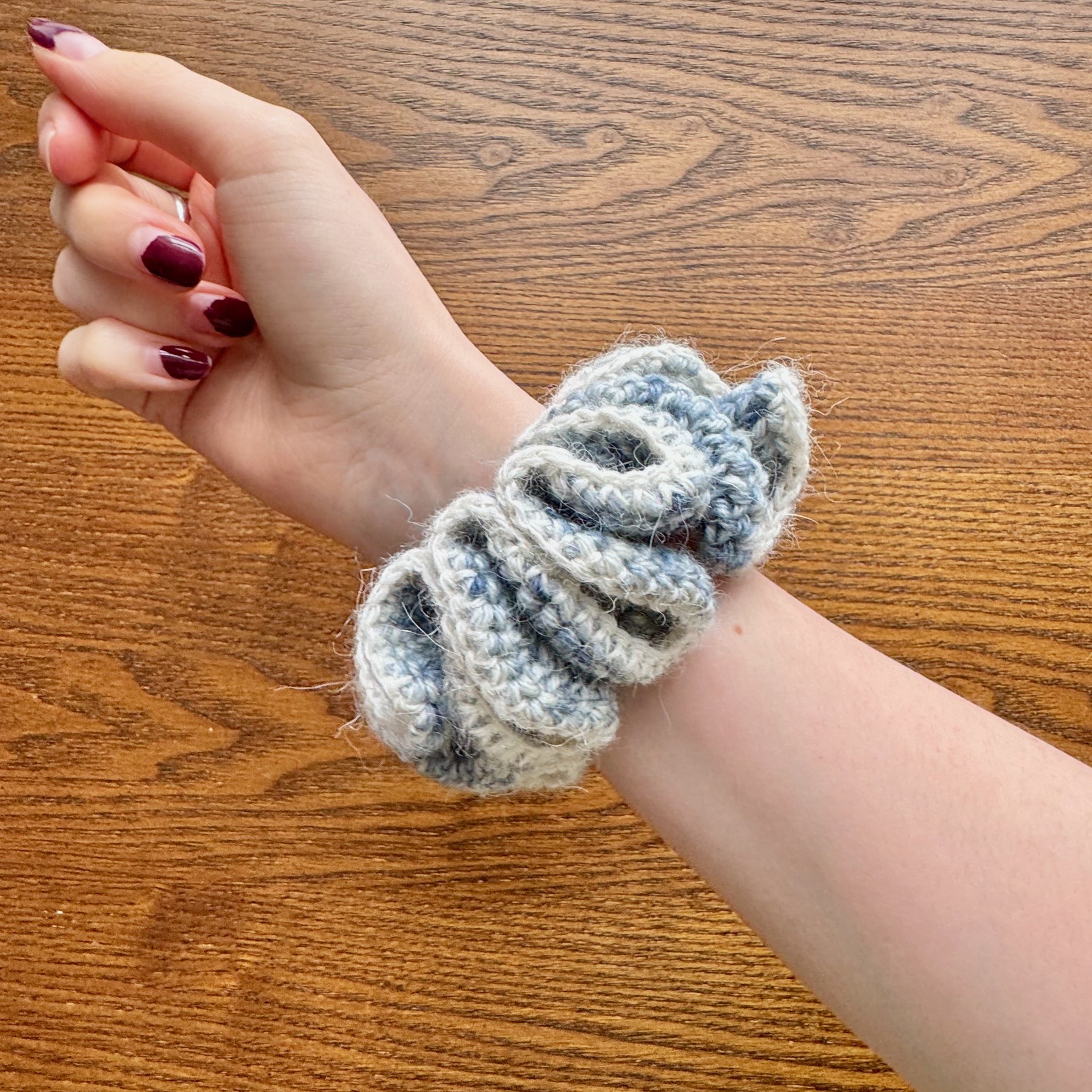 A multi blue woollen crochet ruffle scrunchie that looks like denim with a white alpaca trim being worn on a wrist