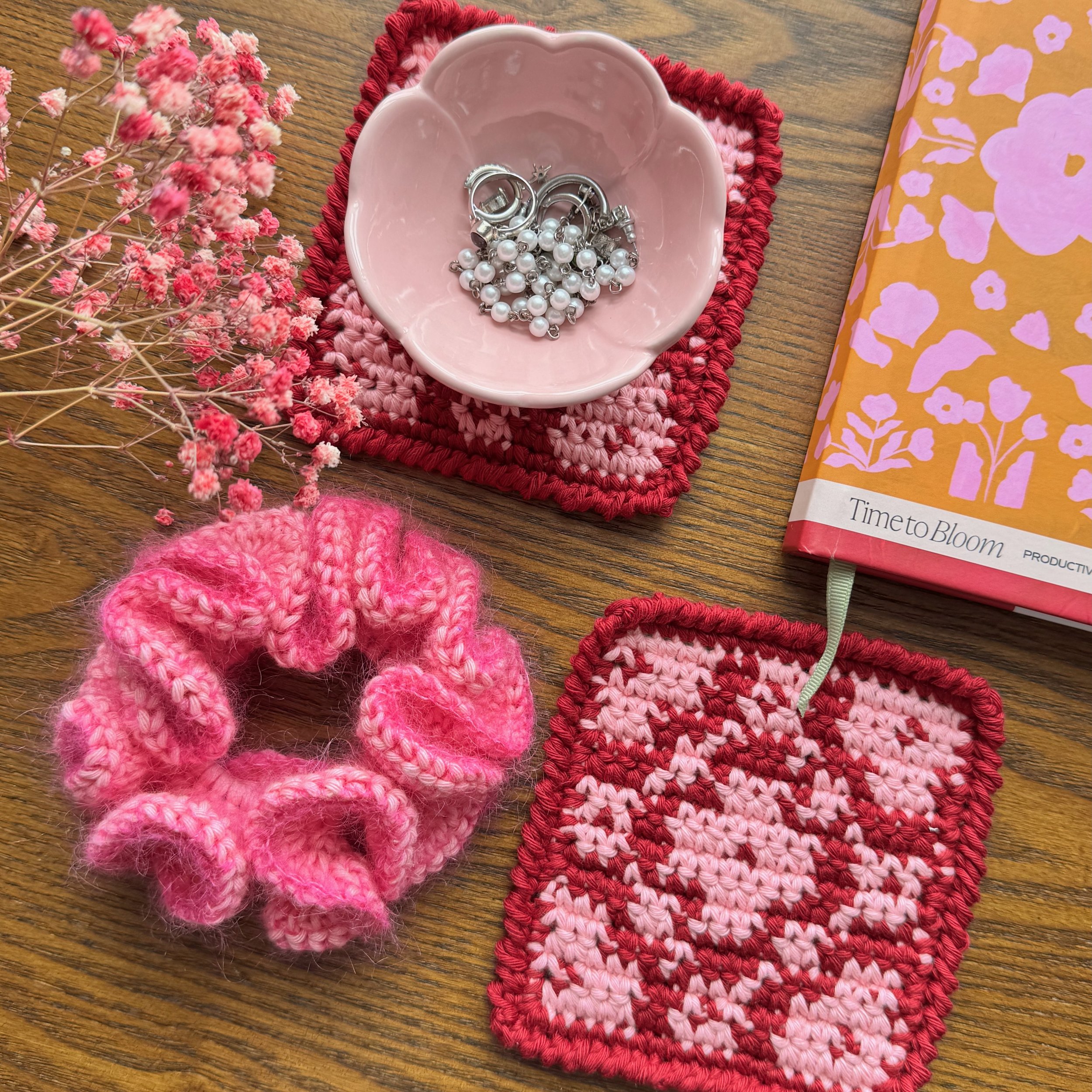 A pink mohair crochet ruffle scrunchie laying flat next to a book, some coasters and a trinket dish