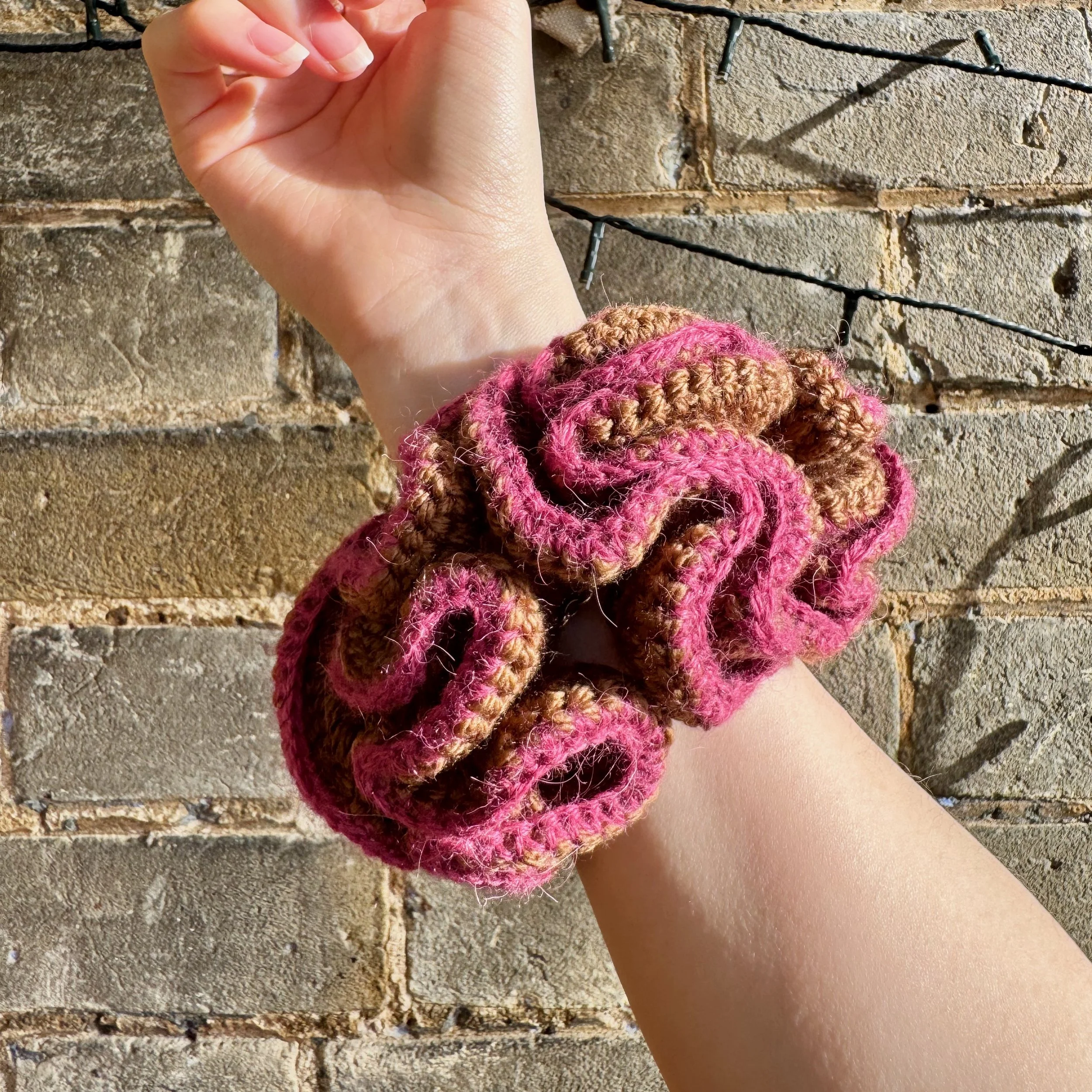 A brown crochet ruffle scrunchie with a pink alpaca trim being worn on a wrist