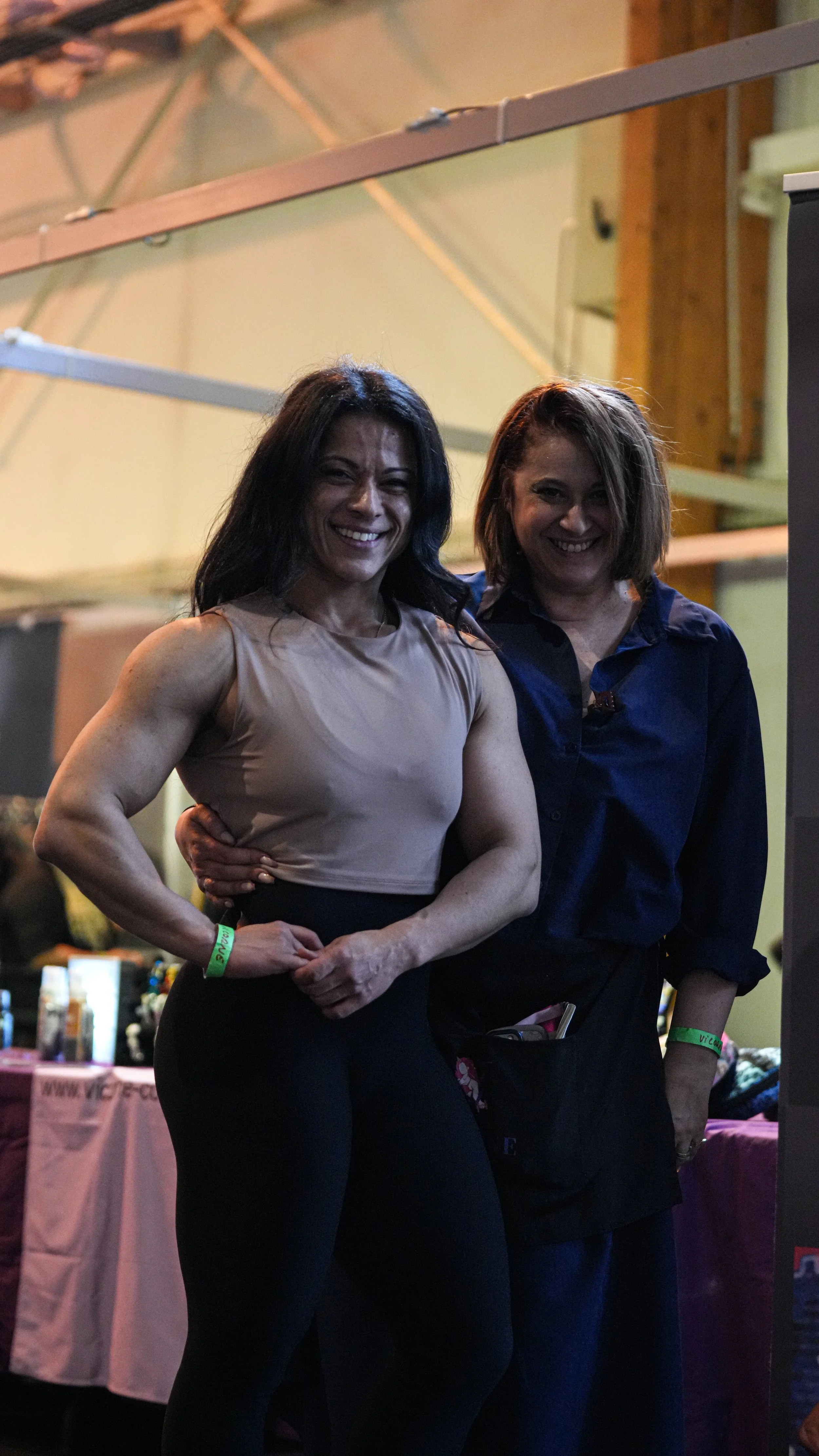 Deux femmes posant, une avec un bustier beige et des muscles visibles, l'autre en chemise bleue. Elles sourient dans un environnement intérieur, probablement lors d'un événement.