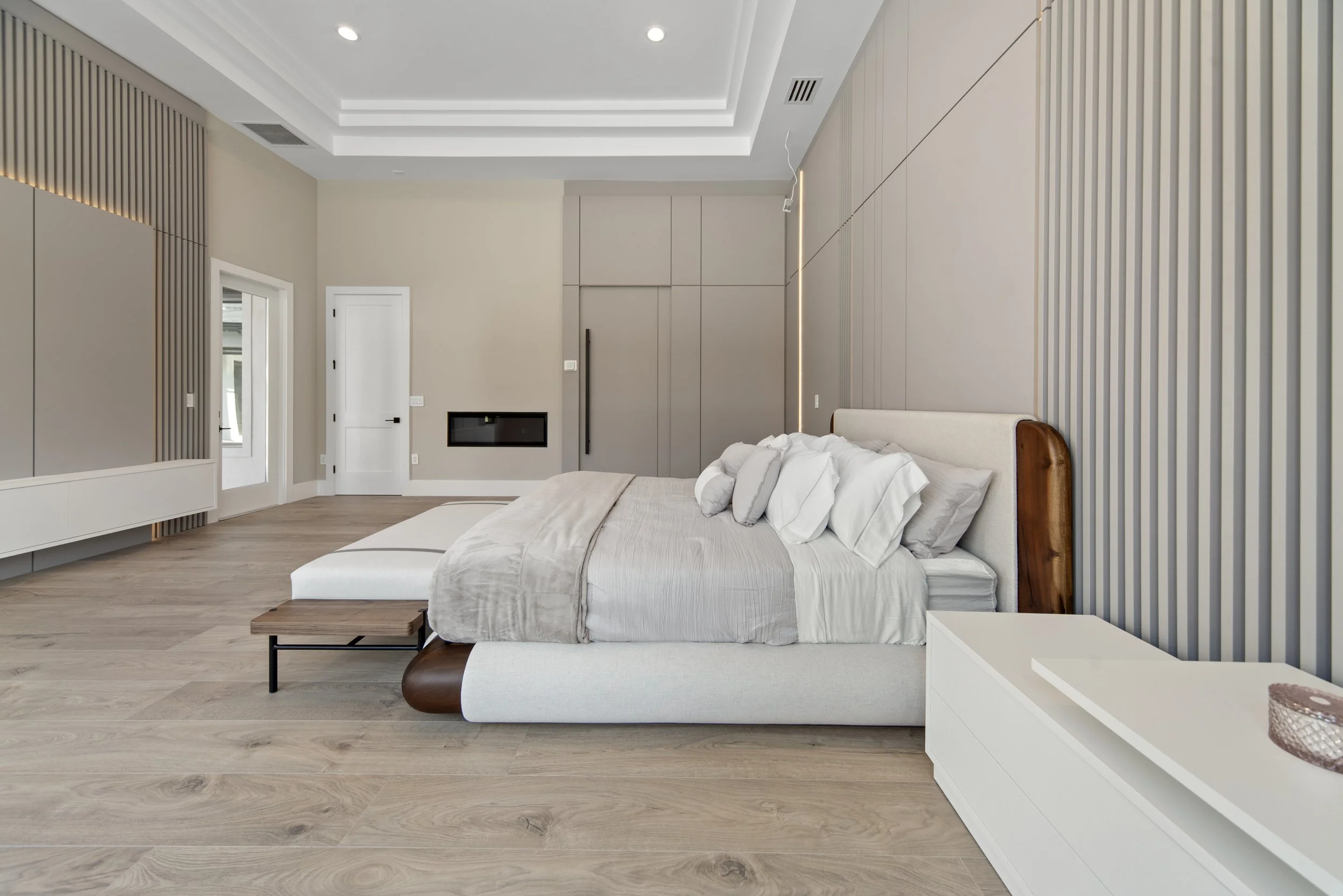 Modern bedroom with beige walls, light wooden flooring, a large bed with cream and tan bedding, and a wooden feature on the headboard. Minimalist furniture and ceiling lights.