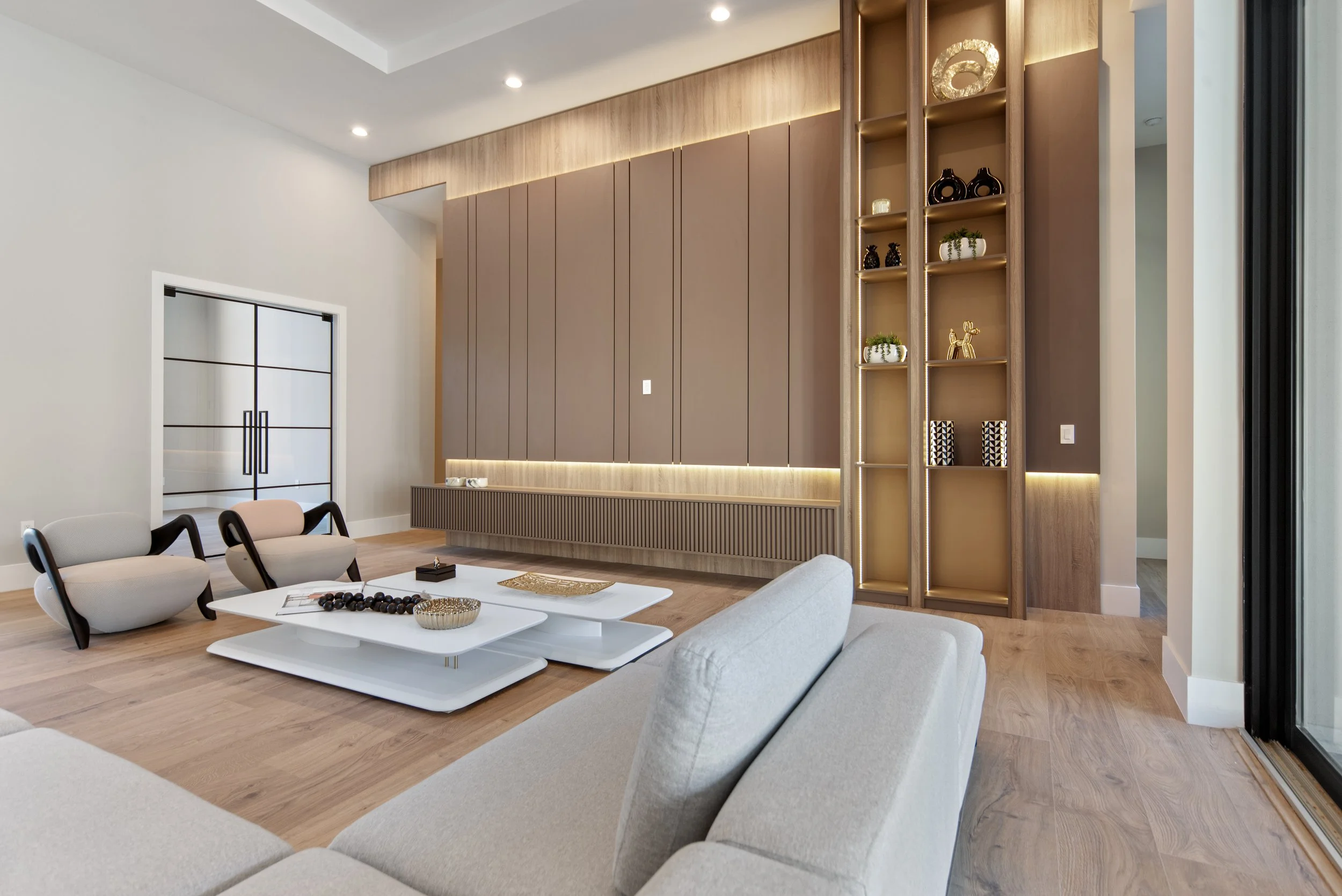 Modern living room with beige sofa, two lounge chairs, a low white coffee table with decor, wooden flooring, a large built-in entertainment unit with decorative shelving displaying vases and sculptures, a sliding glass door, and neutral painted walls