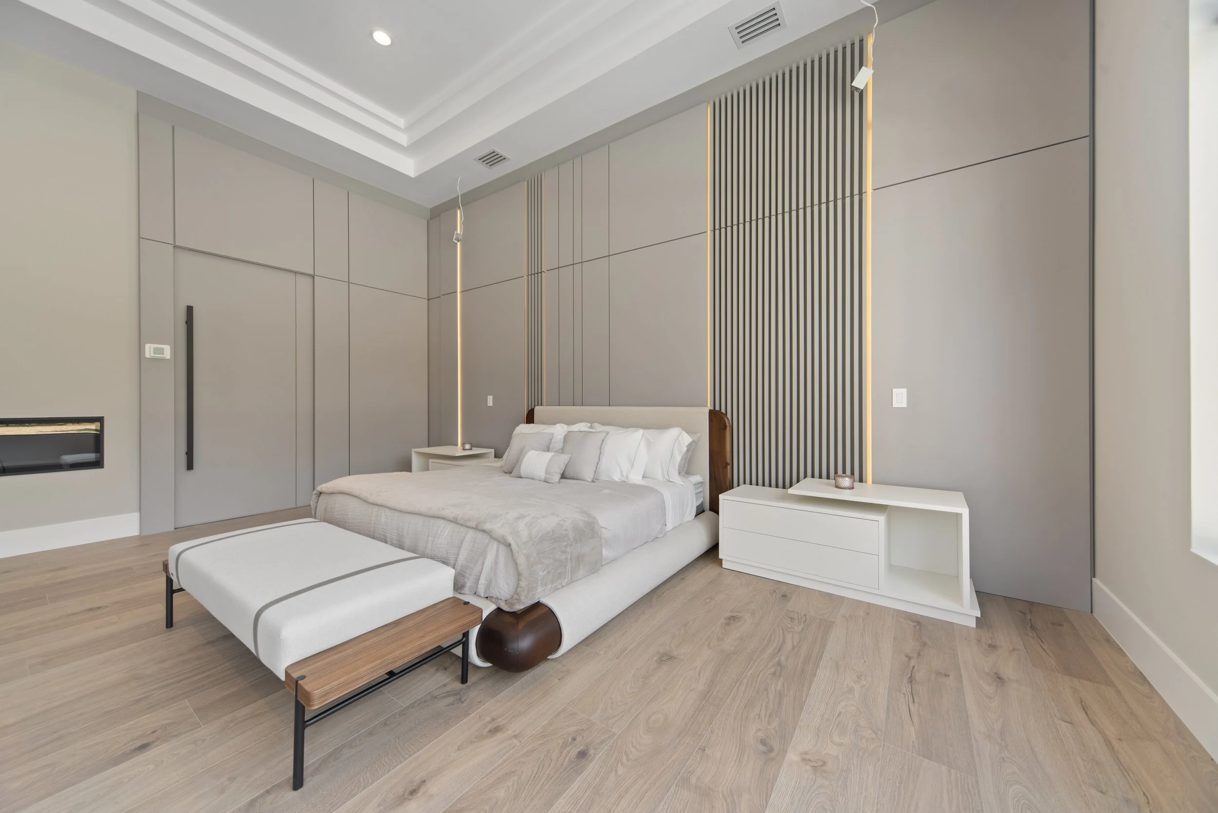 Modern bedroom with a large bed, white and beige bedding, two white nightstands, light wood flooring, a beige wall with vertical decorative panels, and a ceiling with recessed lighting.