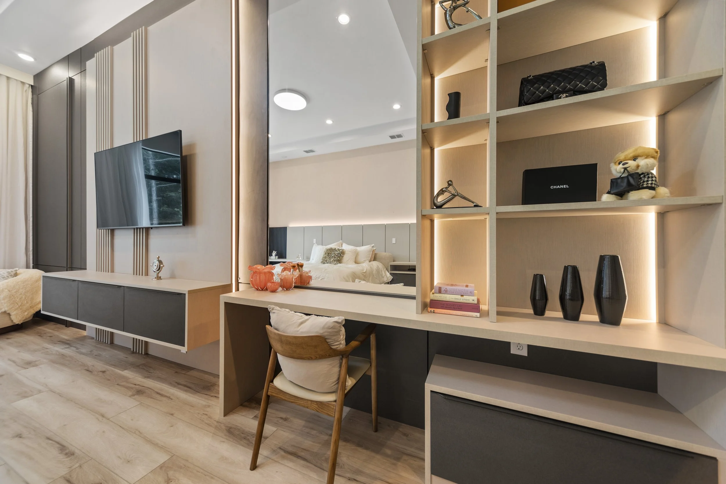 Modern living space with a wall-mounted flat-screen TV, a decorative mirror, a study desk with a chair, and a display shelf with handbags, books, and decorative objects. A bedroom with a bed is visible in the background.