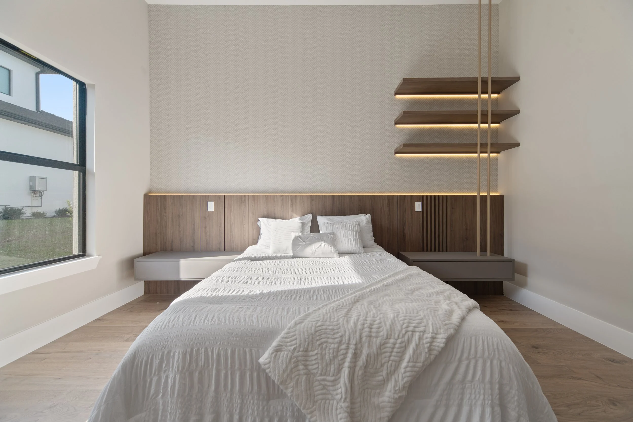 Minimalist bedroom with a large white bed, wooden headboard, floating gray nightstands, and modern wall-mounted shelves with integrated lighting, beside a window showing an outdoor view.