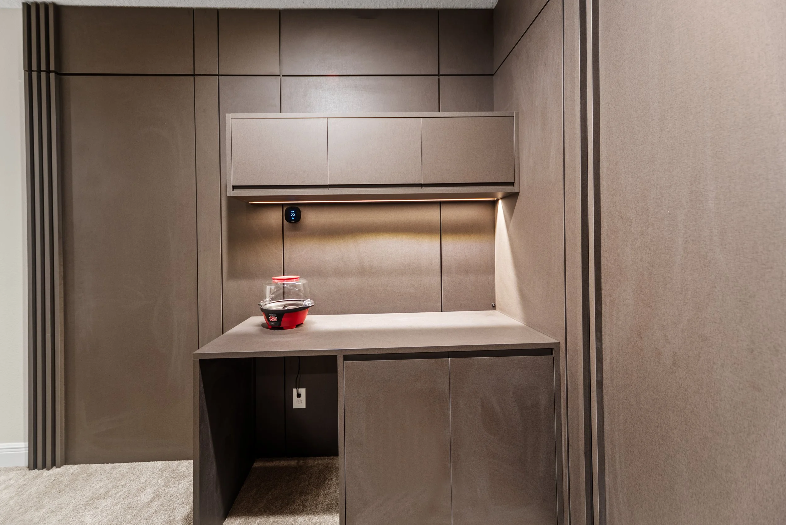 Empty built-in coffee kiosk with a small coffee cup on the countertop, beige wall panels, a digital temperature display, and electrical outlet, in a modern office or conference room.