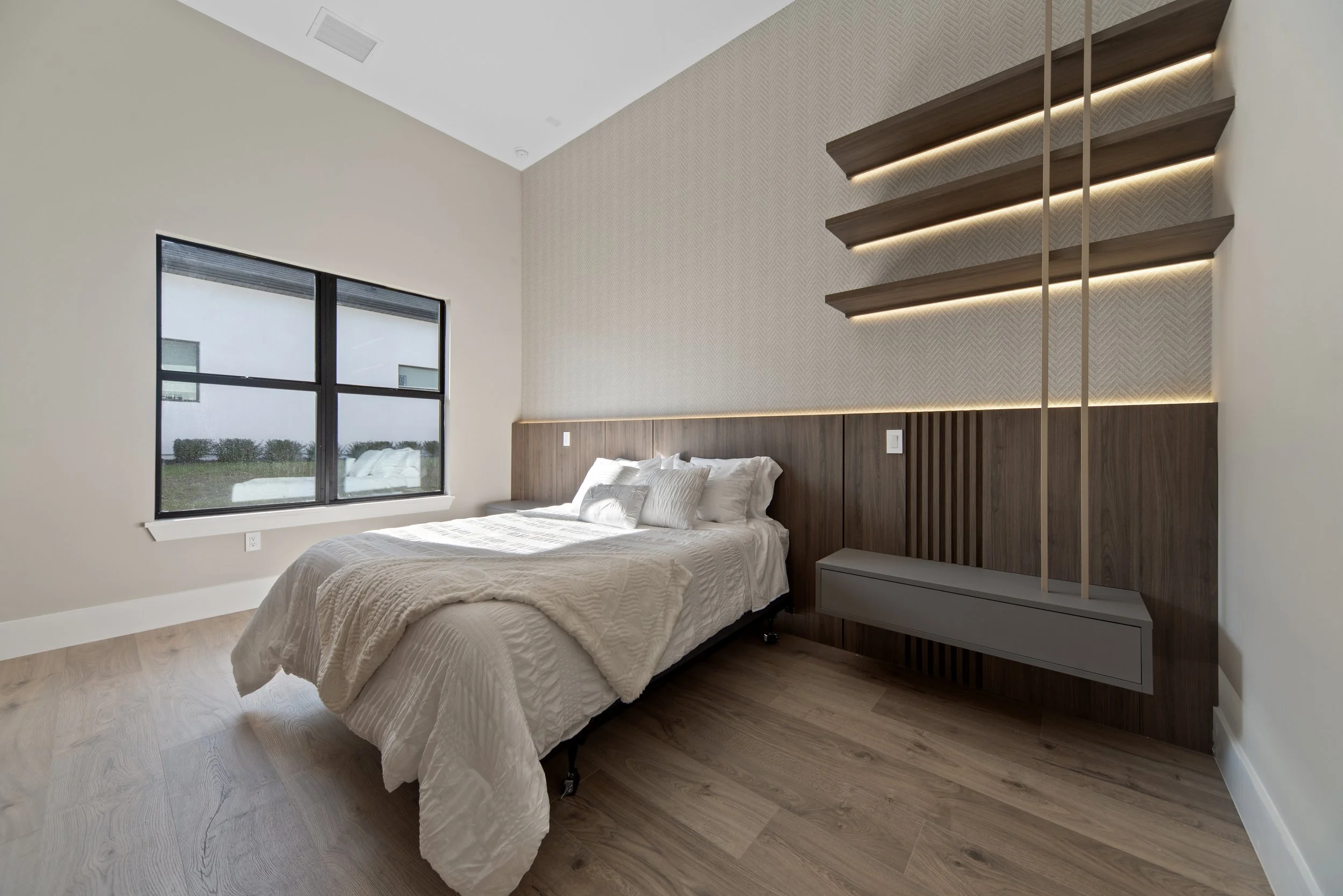 A modern bedroom with a bed, white bedding and pillows, wooden floors, large window showing outdoors, and a textured accent wall with floating wooden shelves and ambient lighting.
