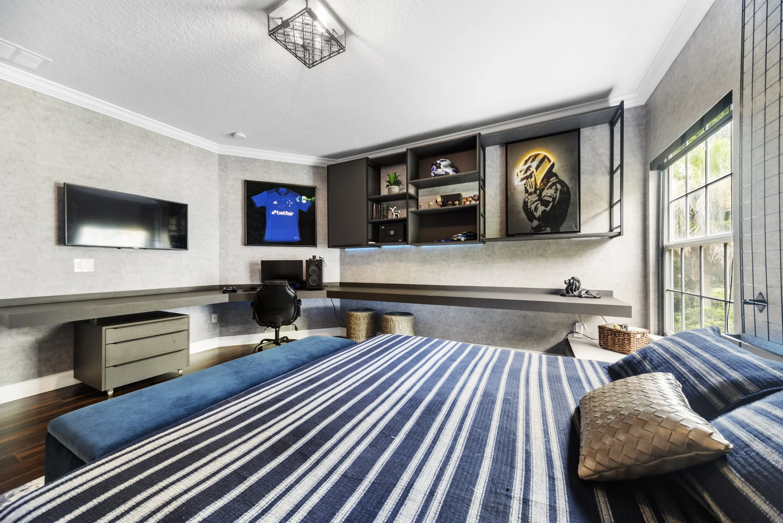 A bedroom with a large bed covered in blue and white striped bedding, a computer desk with a chair, wall-mounted TV, modular shelves with collectibles, sports memorabilia, artwork, and windows letting in natural light.