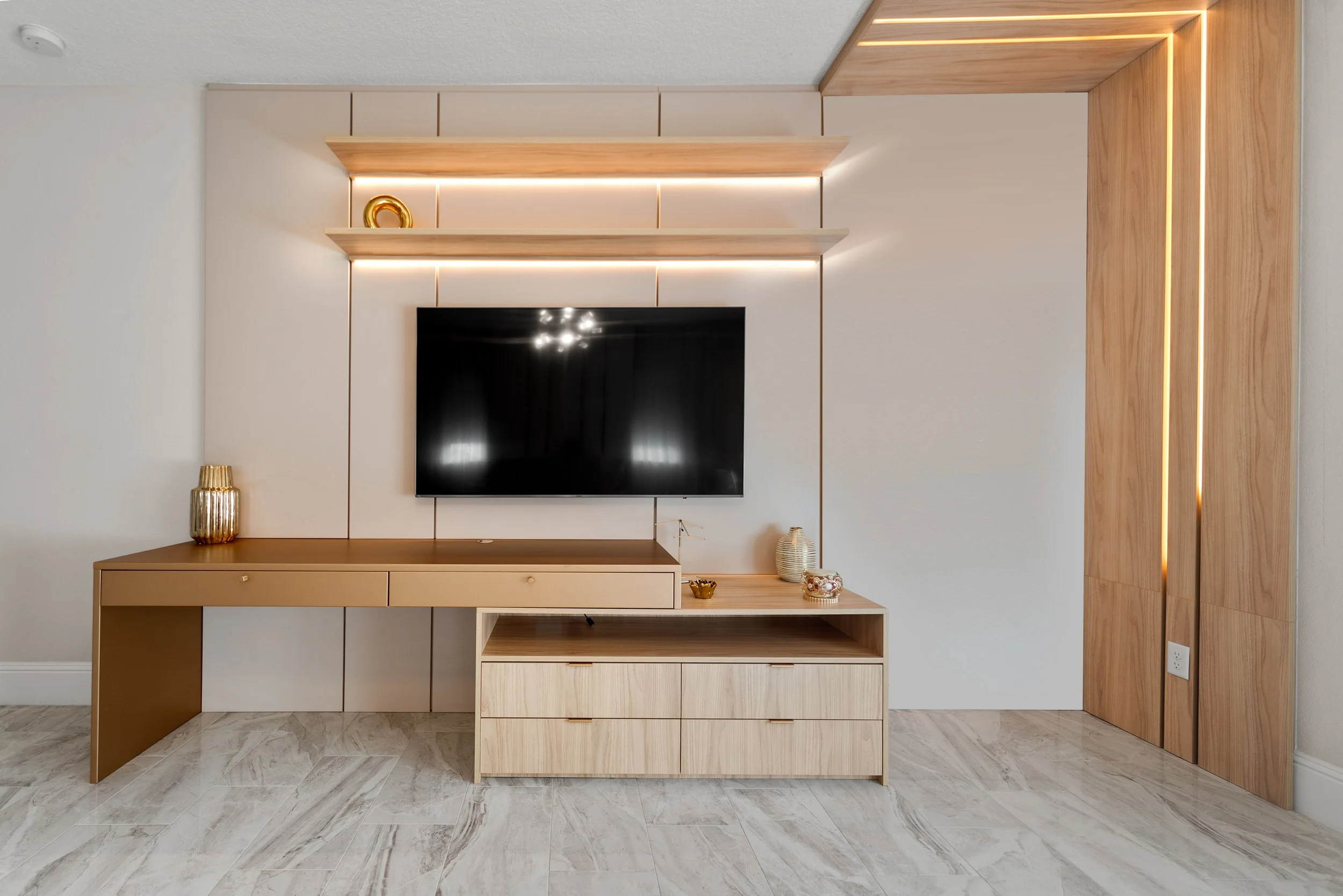 Modern living room entertainment wall with a mounted flat-screen TV, decorative wooden shelves with lighting, and light-colored wooden furniture with decorative vases and accessories.