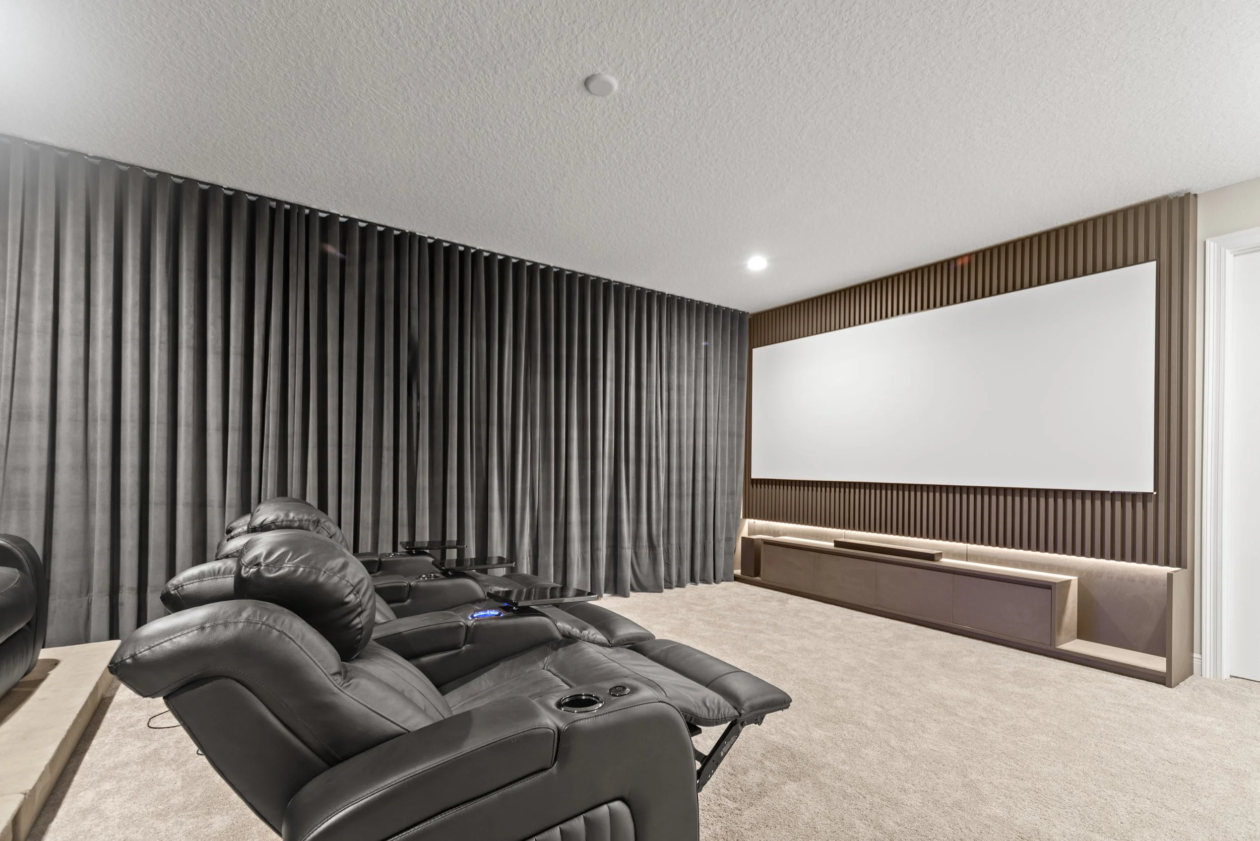 Home theater room with black leather recliner seats facing a large white projection screen, dark curtains on the left side, and a built-in cabinet below the screen.
