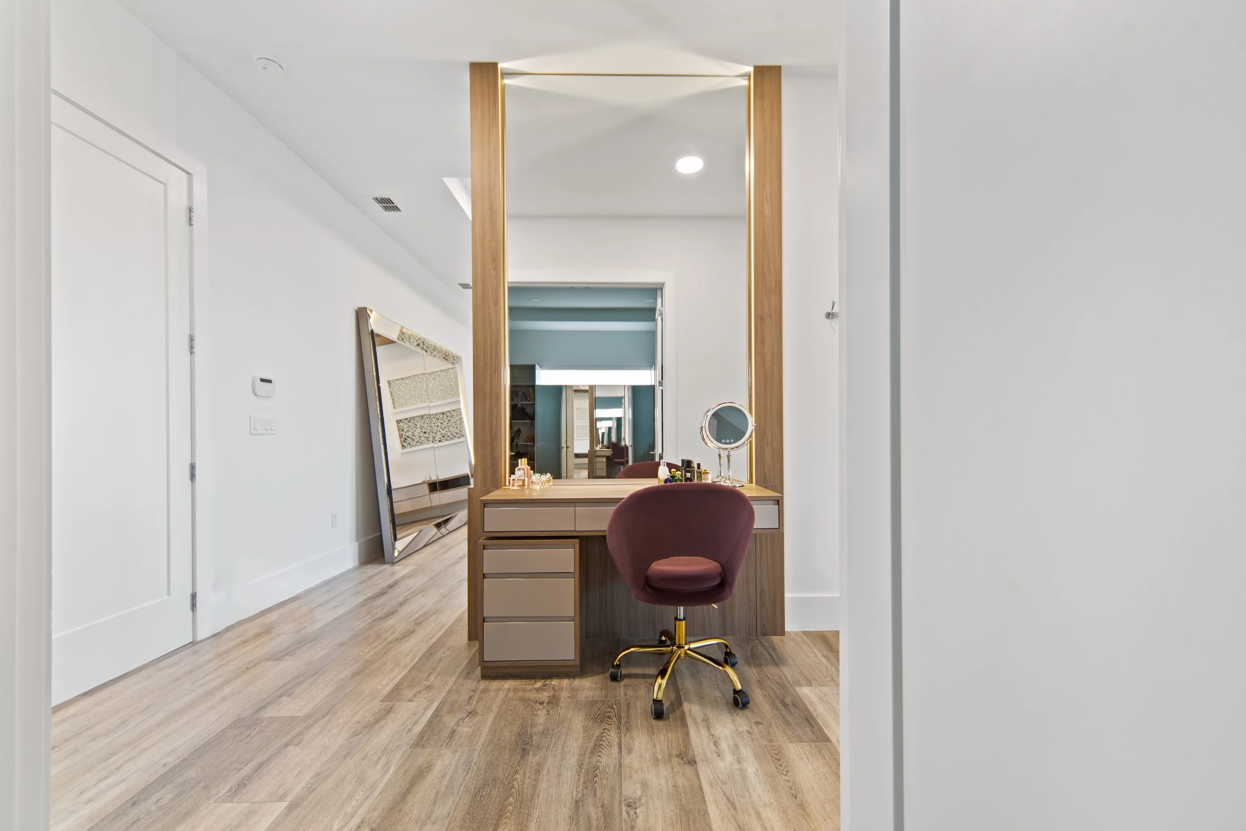 A modern vanity table with a mirror, small makeup accessories, and a cushioned stool in a bright room with wooden flooring.
