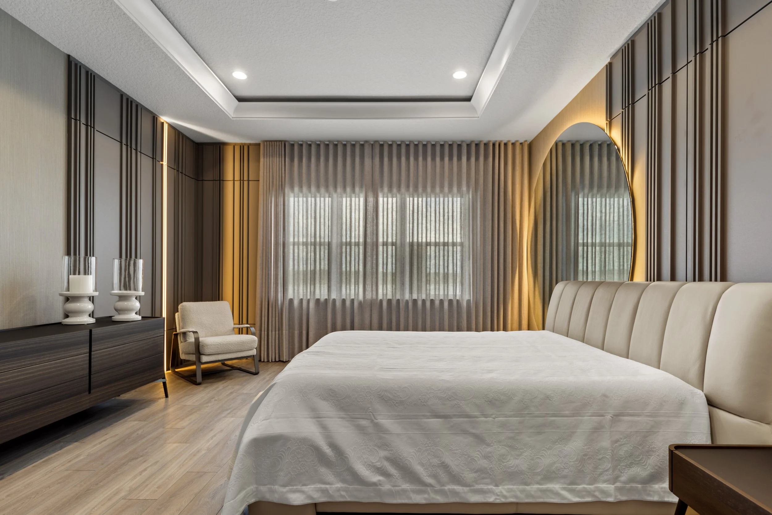 Modern bedroom with a large bed, beige headboard, wooden side table, dresser with candle holders, beige armchair, large window with sheer curtains, ceiling lights, and decorative wall features.